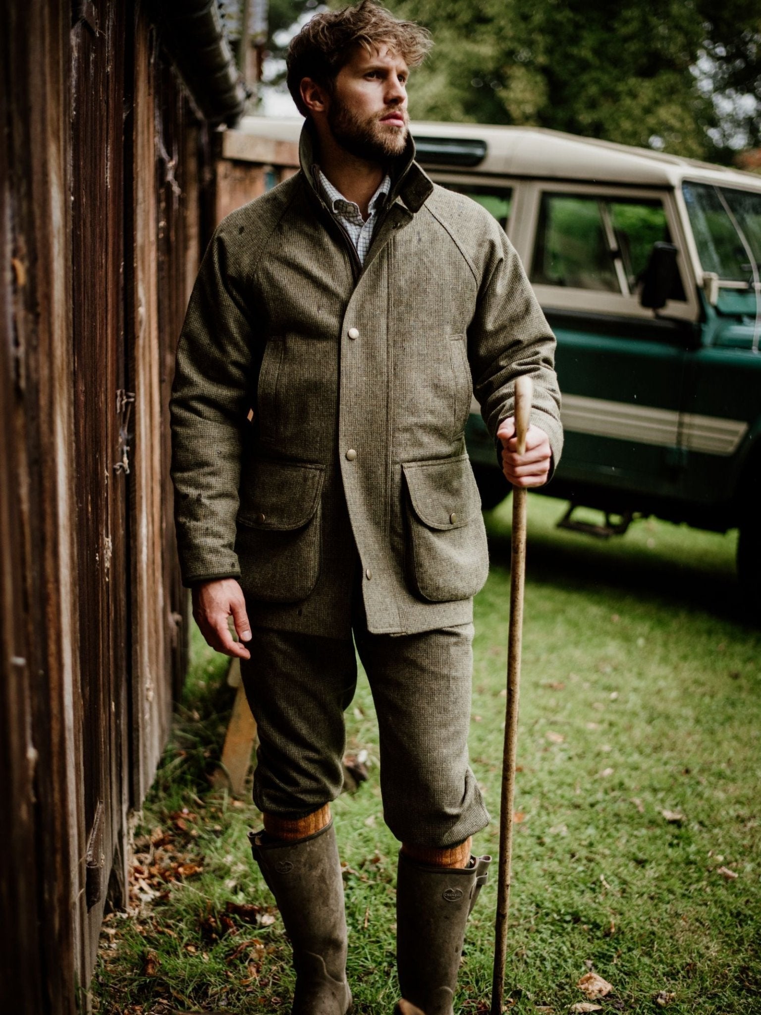 A man wearing Campbells of Beauly Tweed Breeks, a matching green jacket, and brown boots stands on grass by a wooden fence with a walking stick. A vintage green vehicle sits in the background.