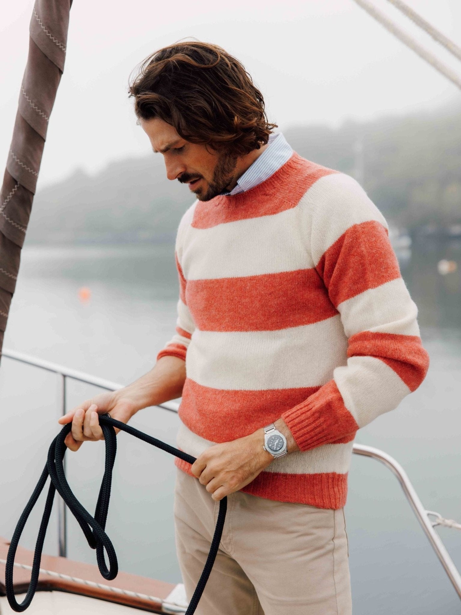 A man wears a Shetland Stripe Crew Jumper by Campbells of Beauly and beige pants as he stands on a boat, holding a black rope and looking down, with misty water and trees in the background.