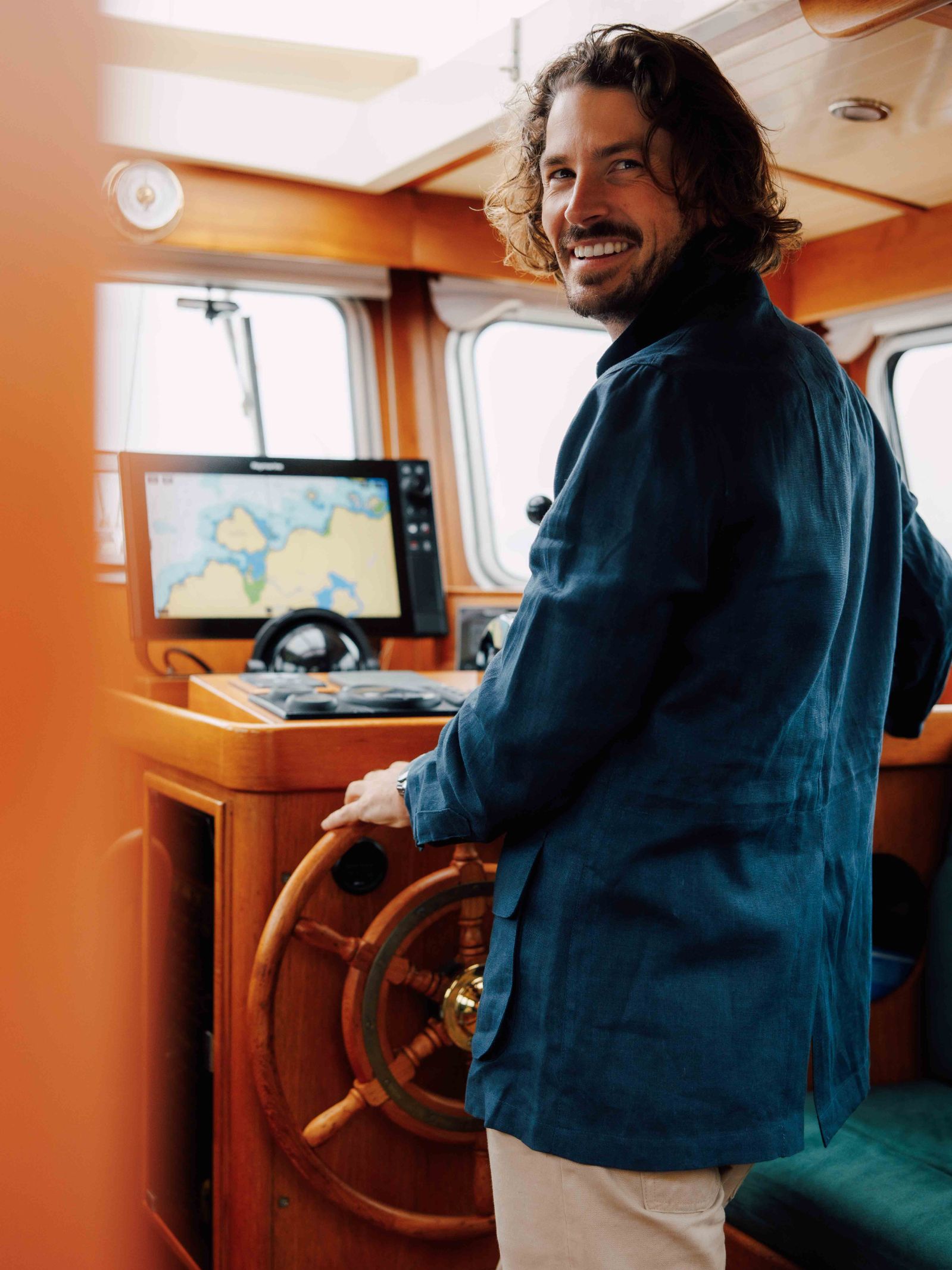 A man with wavy hair and a beard smiles at the wooden wheel of a boat, wearing the Campbells of Beauly Linen Safari Jacket—a stylish casual layer. A nautical map is visible on a screen inside the cabin ahead of him.