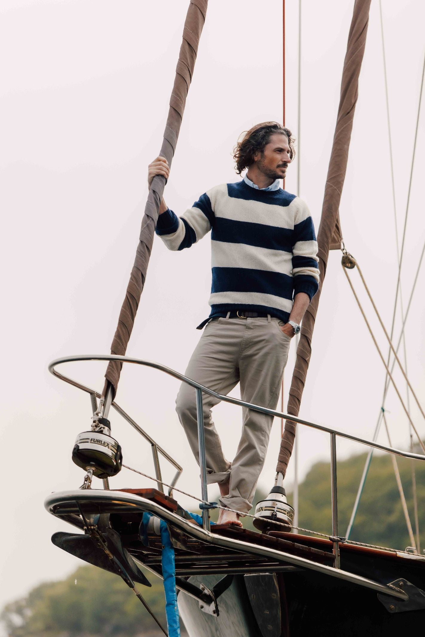 A man in the Campbells of Beauly Shetland Stripe Crew Jumper and light pants stands barefoot at a sailboat’s bow, holding the rigging and gazing ahead, with blurred trees visible in the background.