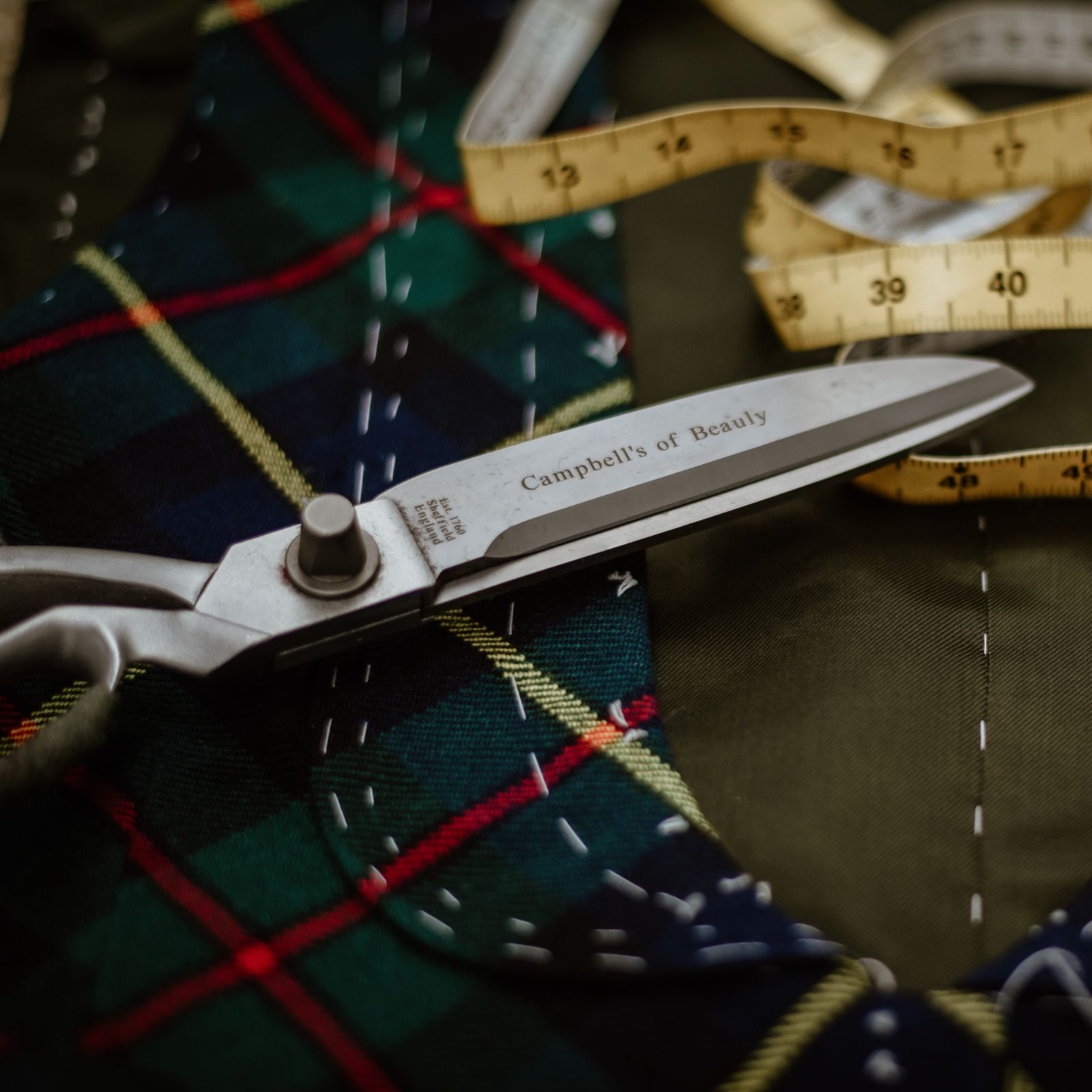 Close-up of fabric with a green, blue, and red plaid pattern, a metal pair of scissors labeled Campbells of Beauly, and a yellow measuring tape, suggesting tailoring or sewing work.