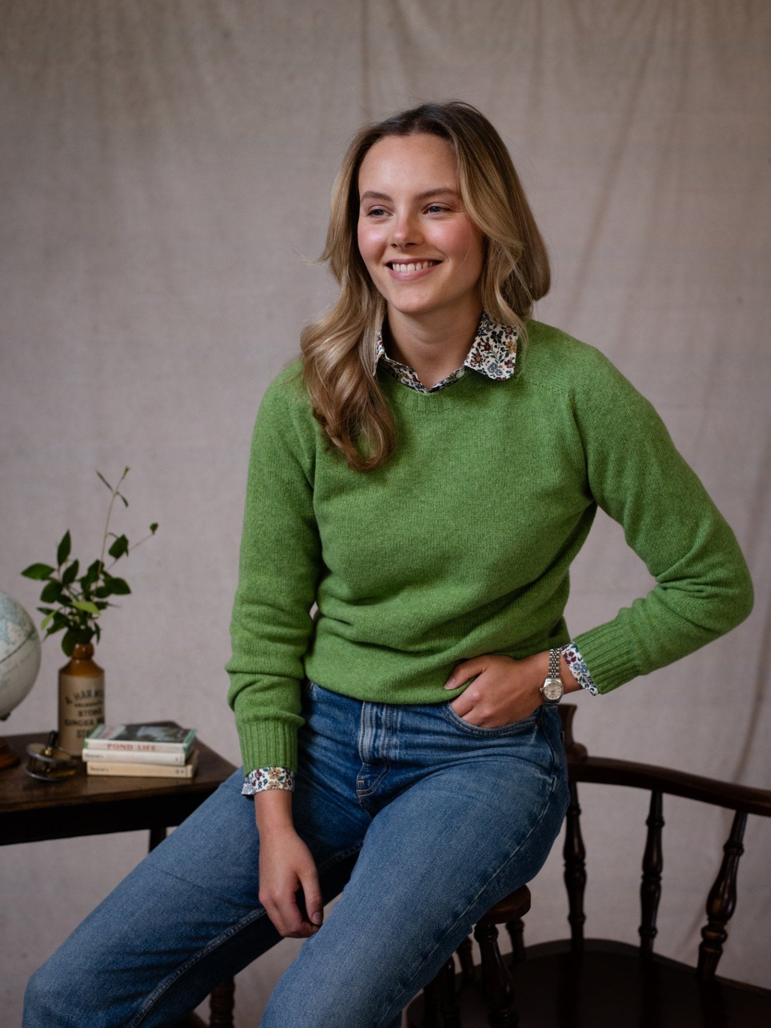A blonde woman, smiling, sits on a wooden chair wearing Campbell's of Beauly's Geelong Lambswool Crew Neck Jumper in green over a floral shirt and blue jeans. Next to her is a small table with books, a globe, leafy stems in a vase, and a candle.