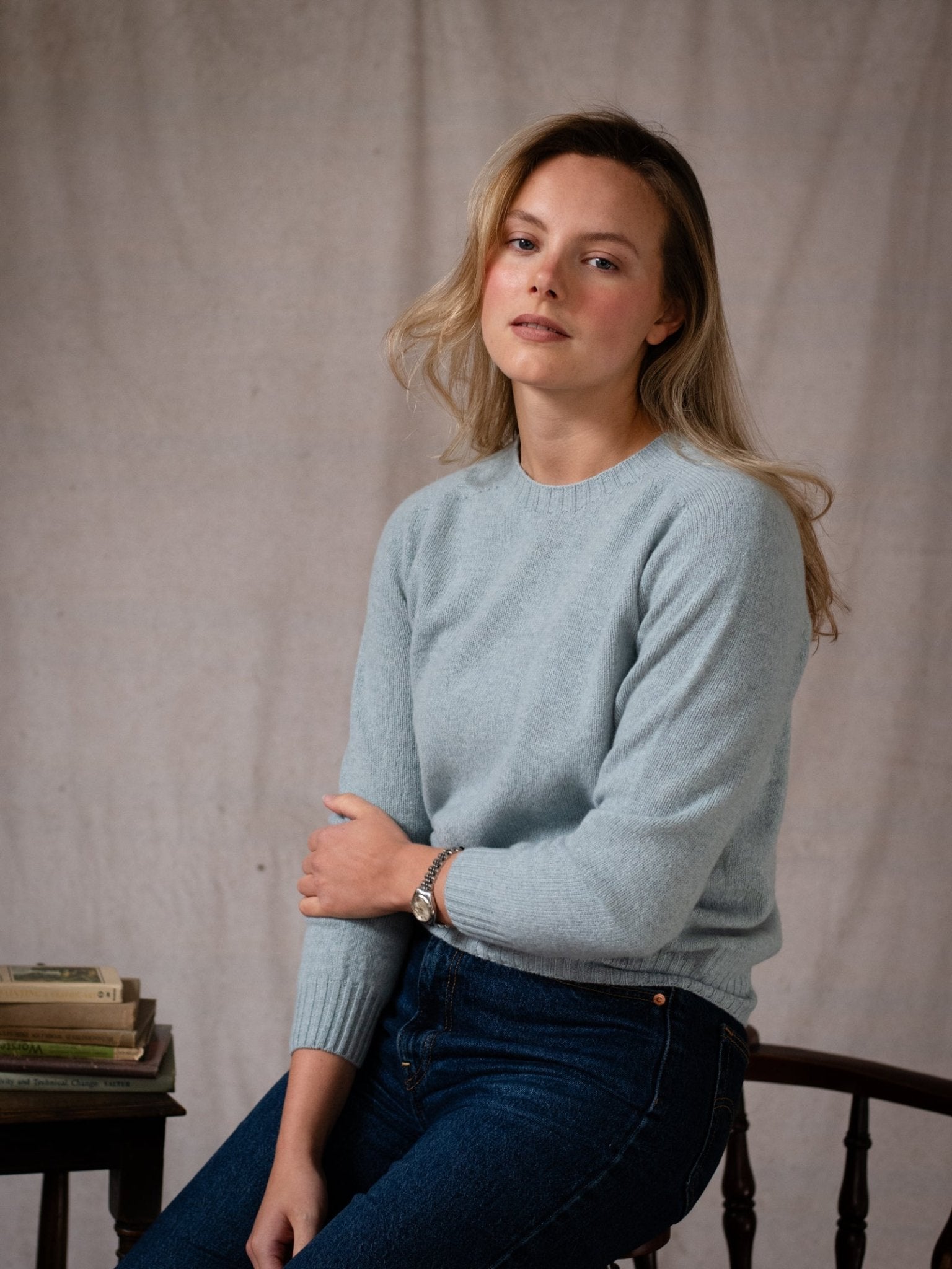 A woman with long blonde hair, wearing the Campbell's of Beauly Geelong Lambswool Crew Neck Jumper in light blue and jeans, sits calmly on a chair beside books against a neutral textured backdrop. Made in Scotland.