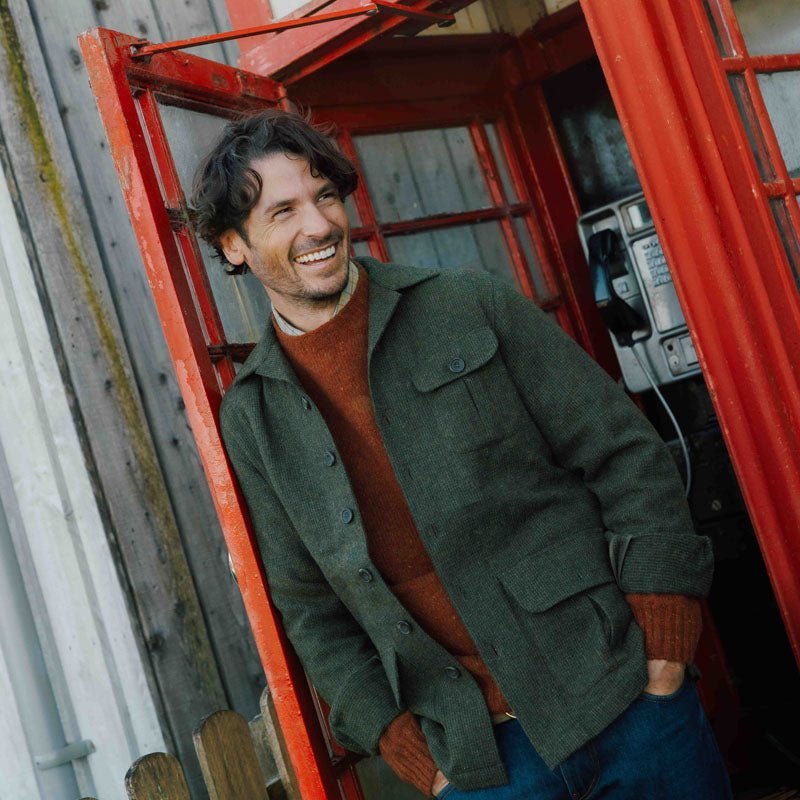 A smiling man with wavy hair stands in a red phone booth, wearing the Auld Stock Casual Tweed Shacket in Green Melange by Campbell's of Beauly over a rust sweater and blue jeans. An old-fashioned payphone is visible inside the booth.