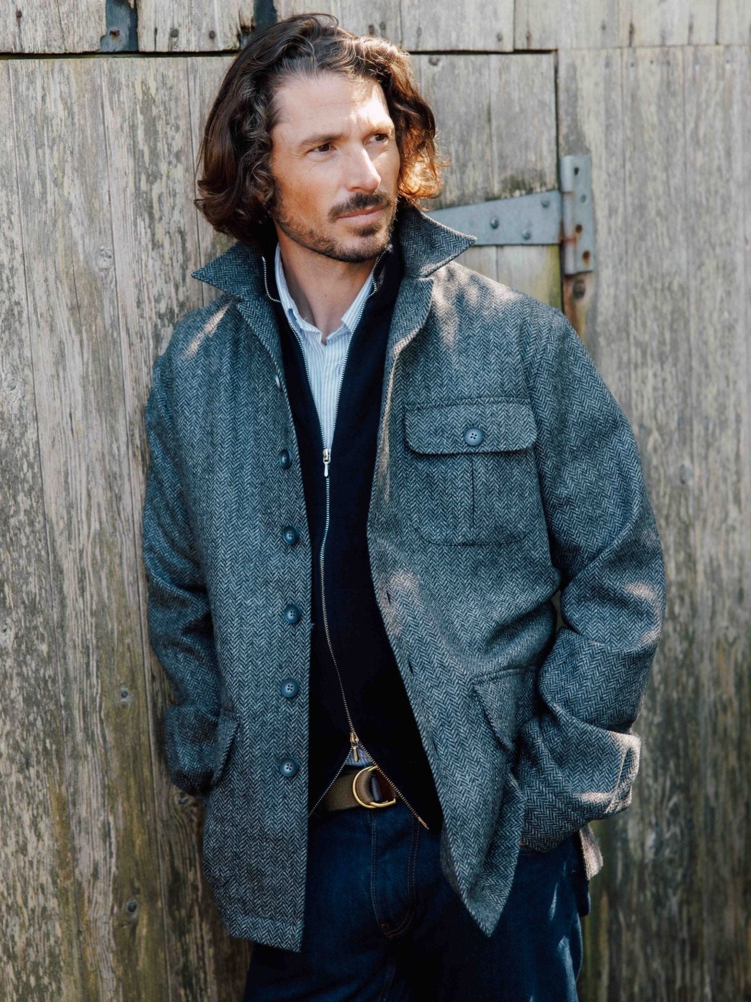 A man with medium-length wavy hair and a beard stands against a weathered wooden door, wearing the Casual Shacket by Campbells of Beauly over a sweater and collared shirt, hands in his pockets as he looks to the side.