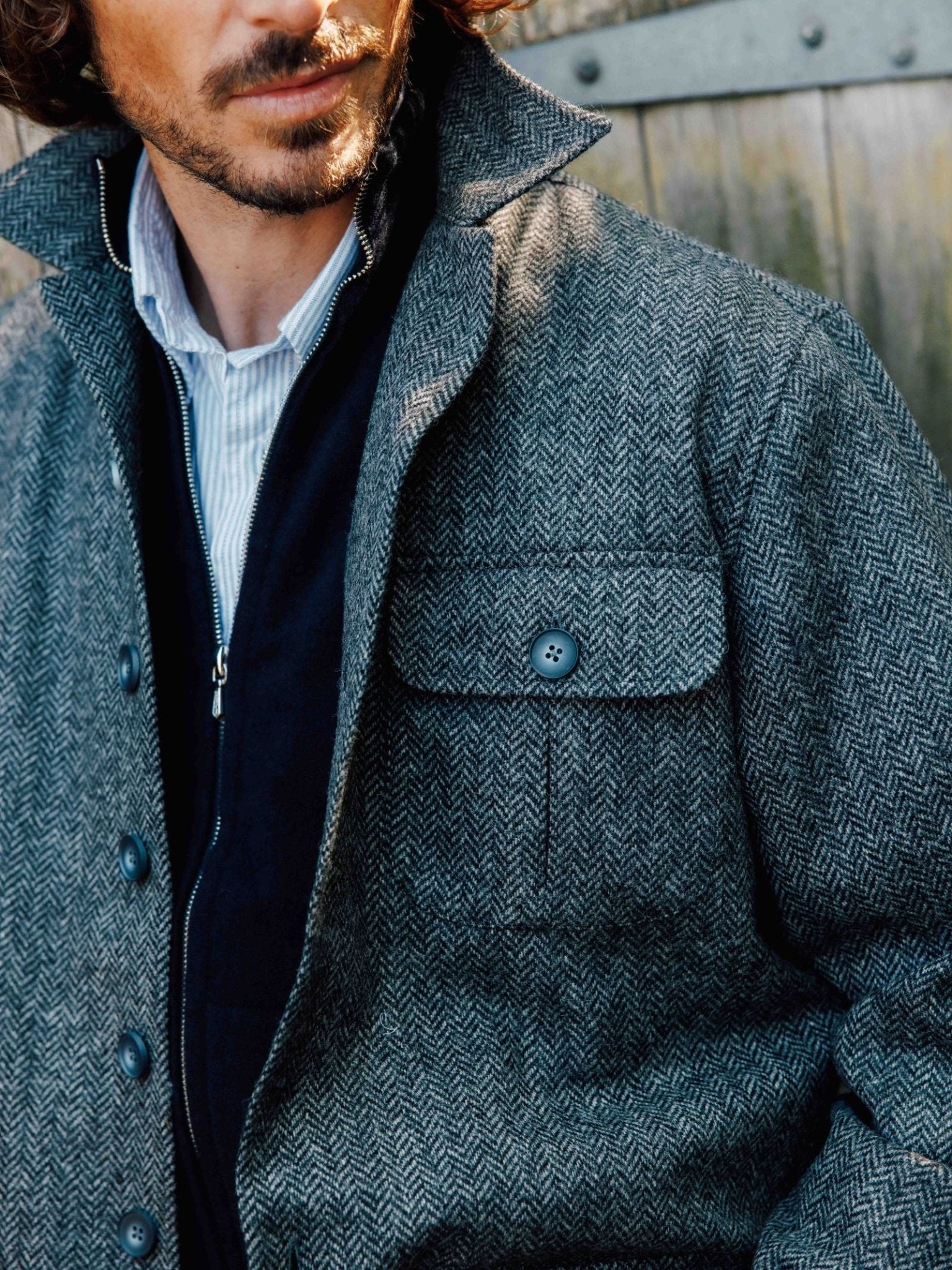 A man stands before a wooden background wearing Campbells of Beauly Casual Shacket—a gray British tweed herringbone piece with a buttoned chest pocket, layered over a dark zip-up and light blue shirt, channeling effortless traditional style.