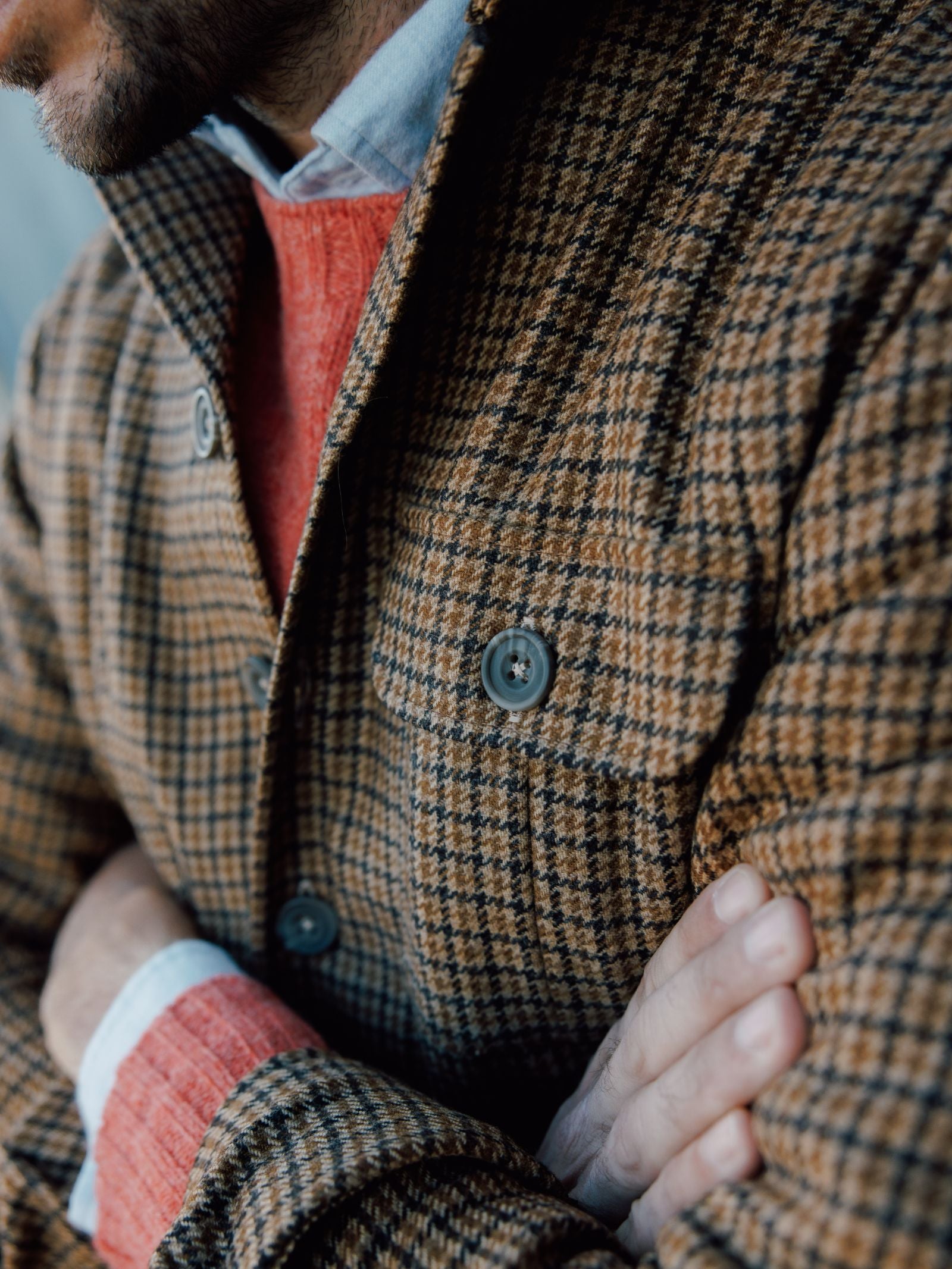 A close-up shows a man in a Campbells of Beauly Casual Shacket, worn over an orange-red sweater and white collared shirt. Only his torso, folded arms, and part of his face are visible.
