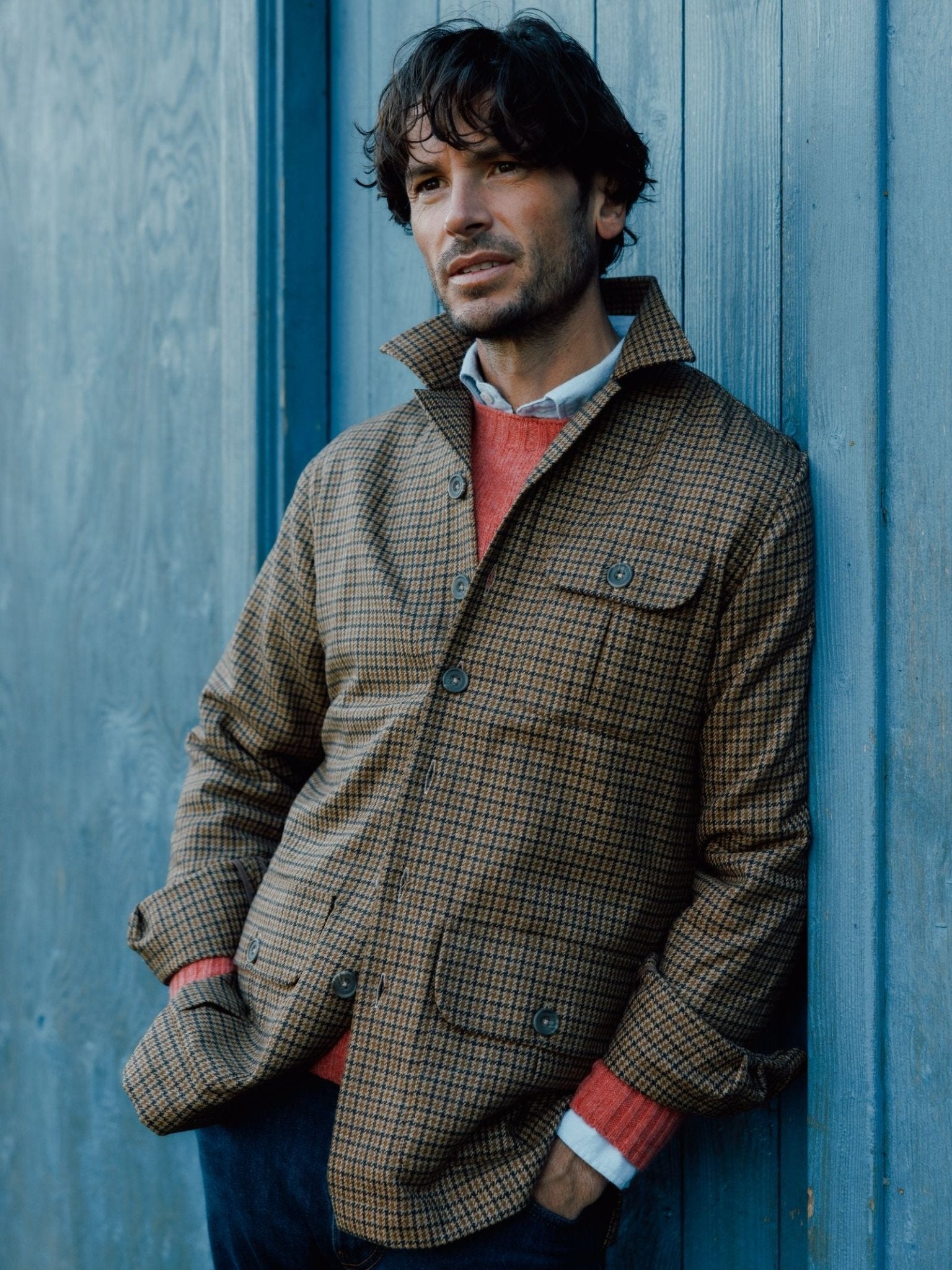 A man with wavy brown hair thoughtfully leans against a blue wooden wall, wearing the Campbells of Beauly Casual Shacket over a red sweater and white collared shirt.