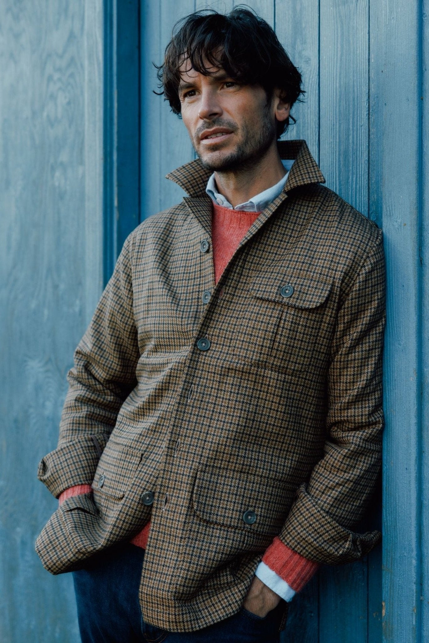 A man with wavy brown hair thoughtfully leans against a blue wooden wall, wearing the Campbells of Beauly Casual Shacket over a red sweater and white collared shirt.
