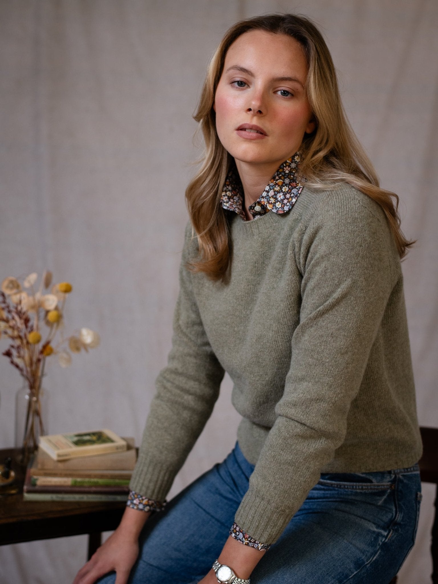 A woman with long blonde hair wears the Campbell's of Beauly Geelong Lambswool Crew Neck Jumper over a floral shirt and blue jeans, sitting indoors beside a table with dried flowers and books against a neutral background.