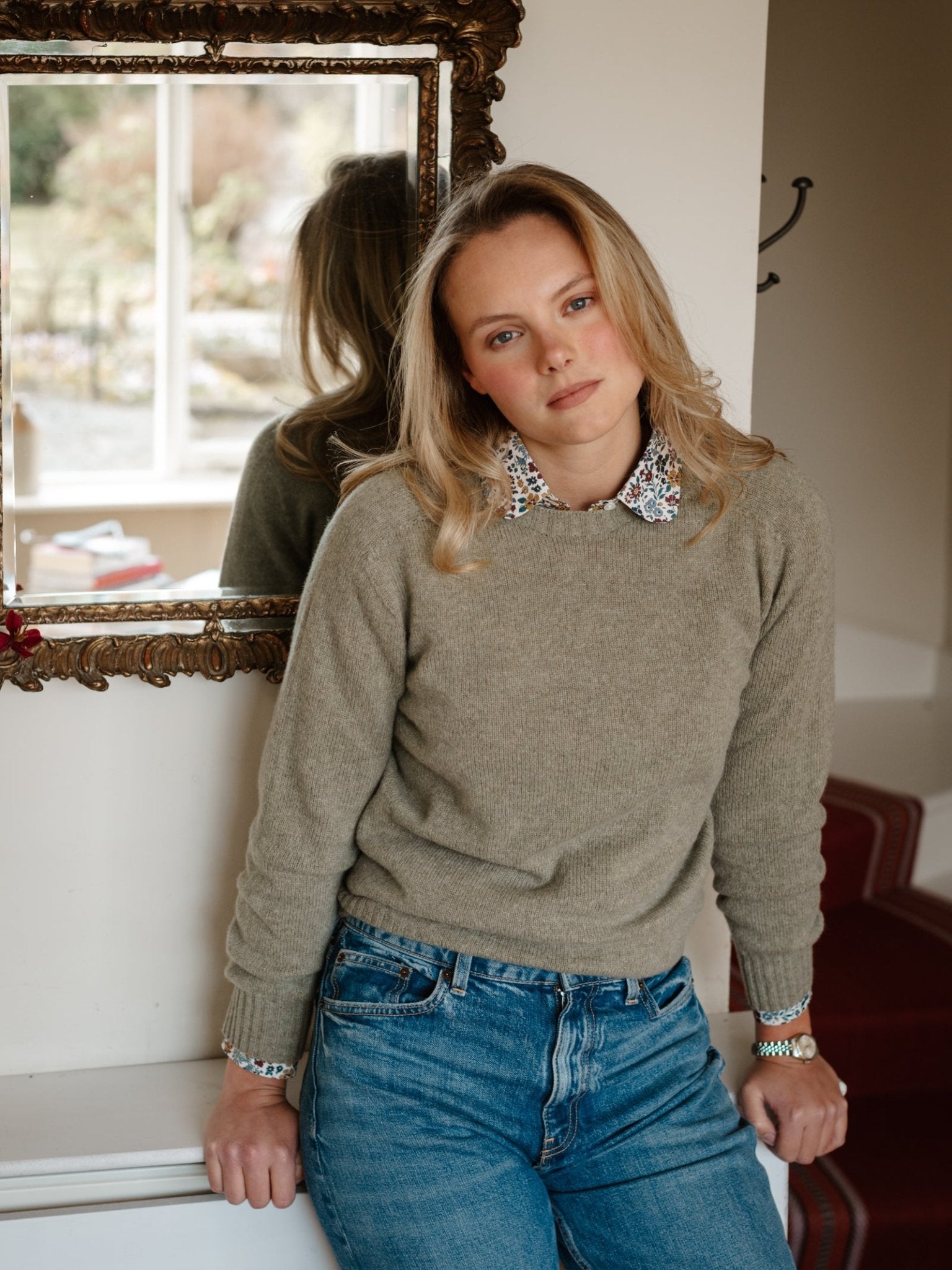 A woman with blonde hair wears a Campbell's of Beauly Geelong Lambswool Crew Neck Jumper over a floral-collared shirt and blue jeans, leaning on a white table before an ornate mirror in a cozy, well-lit room.