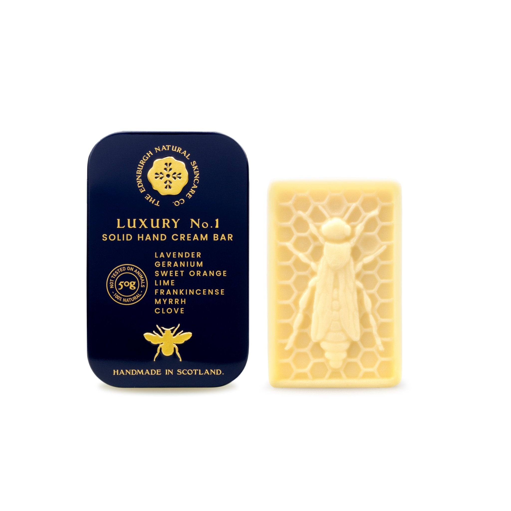 A dark blue tin of The Edinburgh Natural Skincare Company’s Luxury No. 1 Solid Hand Cream Bar sits beside a cream bar embossed with a bee and honeycomb pattern, featuring lavender, geranium, and sweet orange for luxurious natural skincare.
