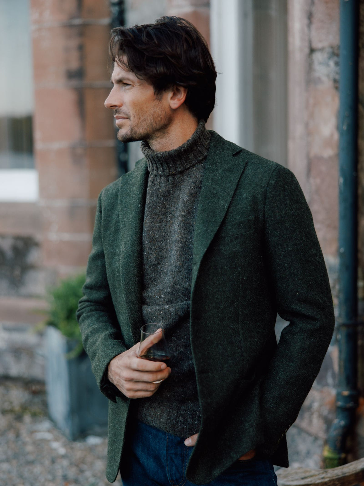 A man with brown hair stands outdoors holding a drink, wearing a dark green Harris Tweed Jacket by Campbell's of Beauly over a gray turtleneck and blue jeans. He looks left, with a brick wall and window in the background.