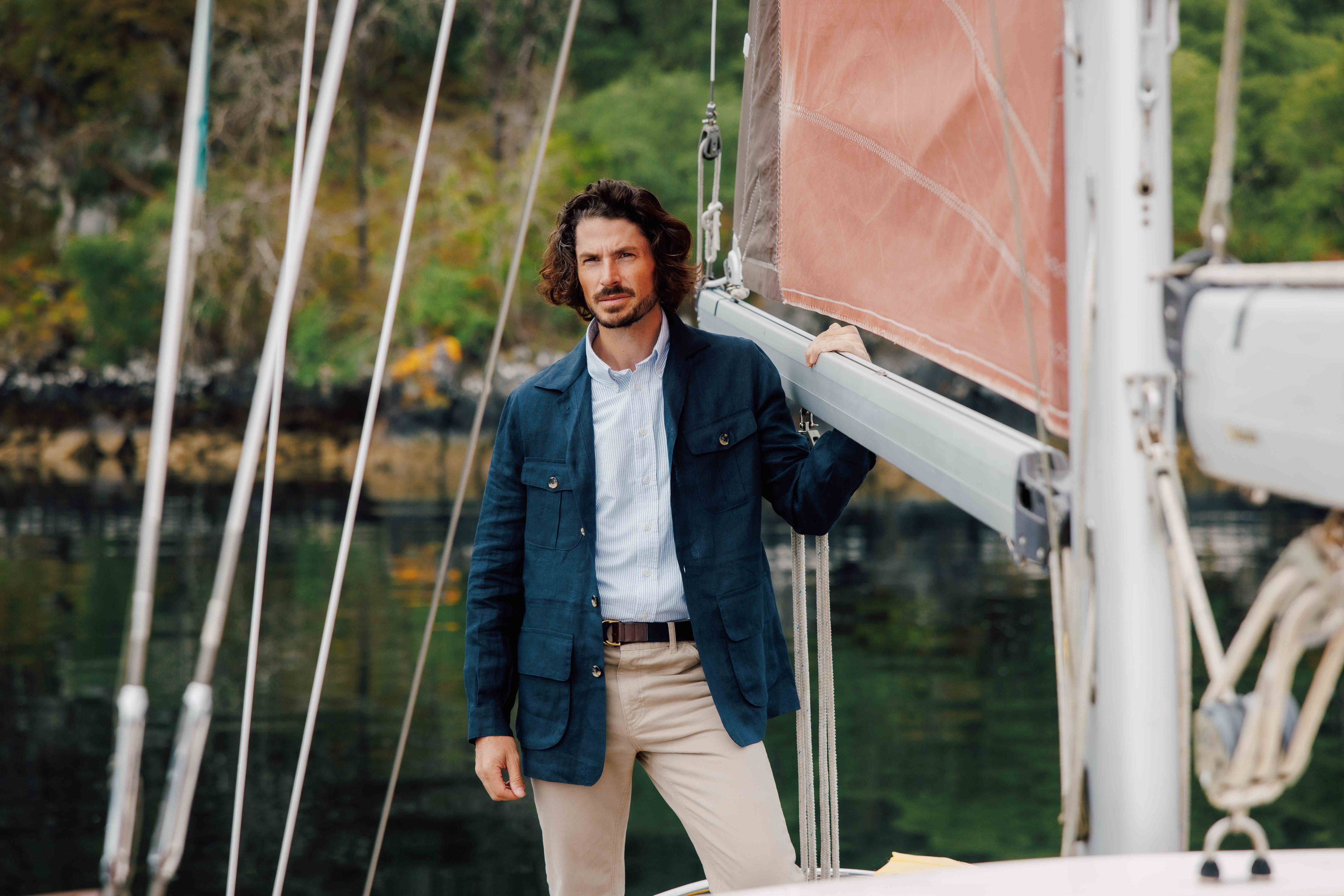 A man with wavy hair, wearing a blue blazer over a light shirt and beige pants, stands on a sailboat holding onto the boom, with trees and water visible in the background.