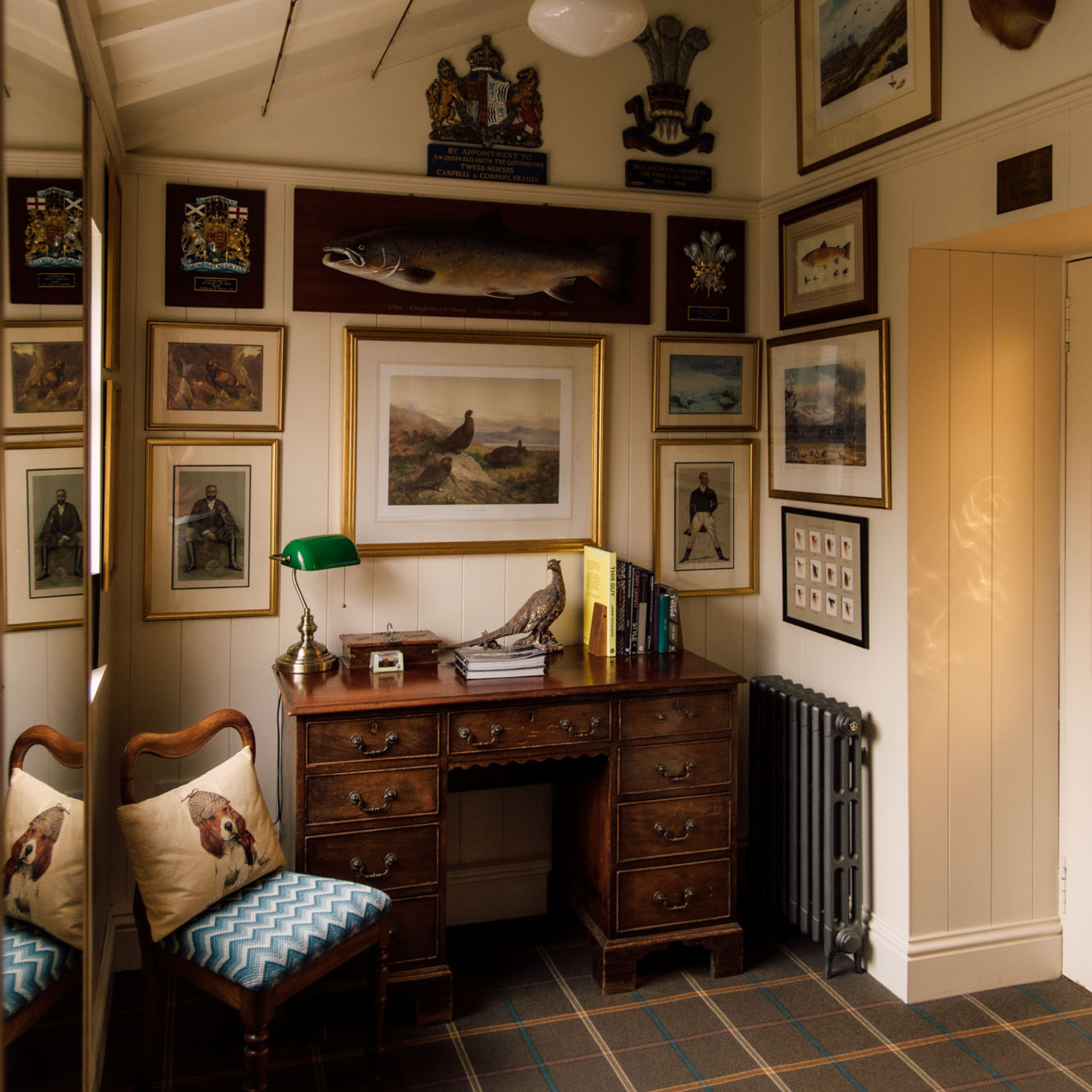 A cozy study corner with a wooden desk, vintage chair, books, lamp, bird sculpture, and framed art and crests covering cream-colored walls. A mounted fish is displayed above the desk. Light filters through from the right.