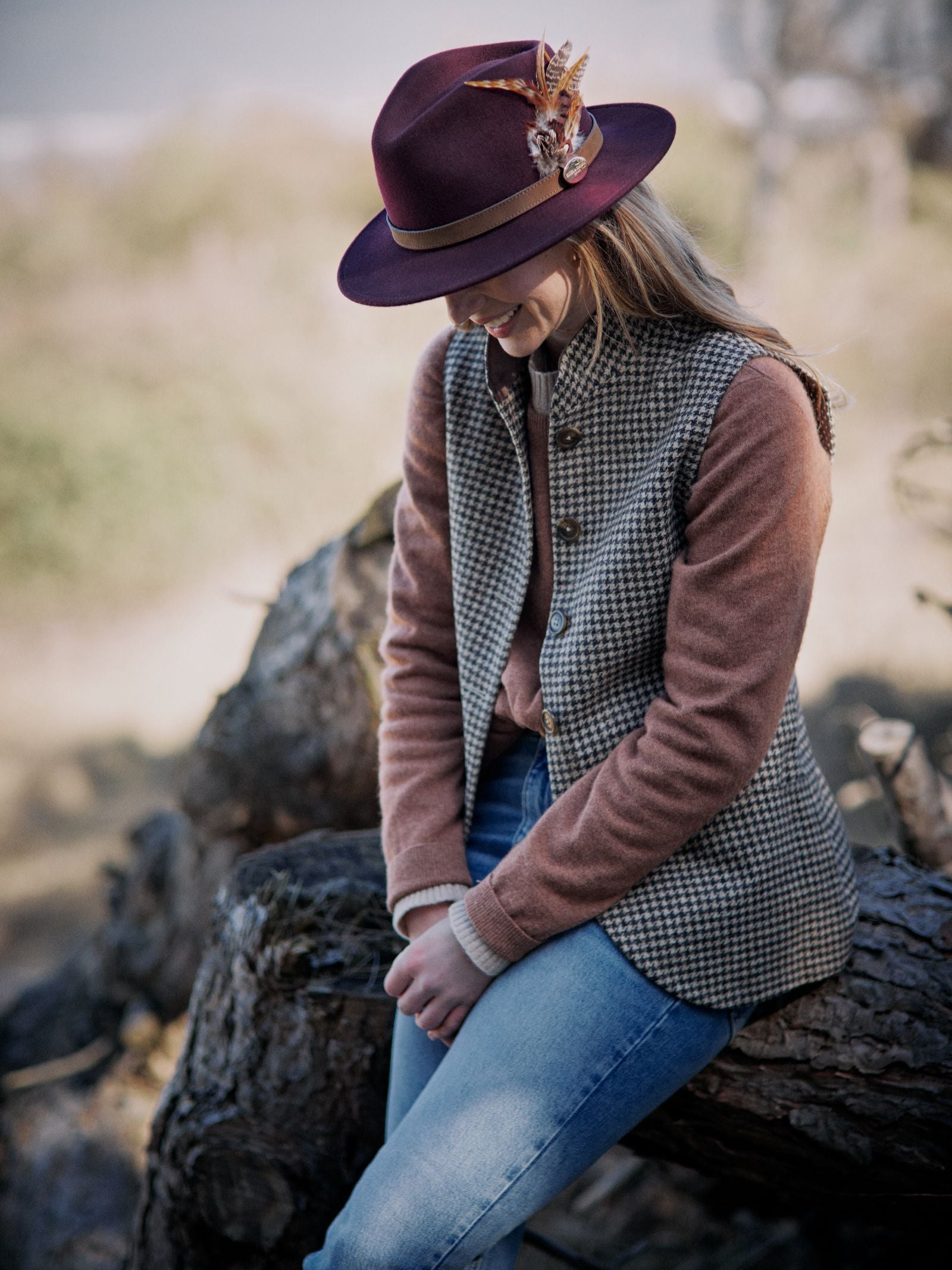 A woman in a maroon Hicks & Brown Gamebird Fedora sits outdoors on a log, wearing feather decorations, a checked vest, pink sweater, and blue jeans, looking down with hands clasped in her lap.