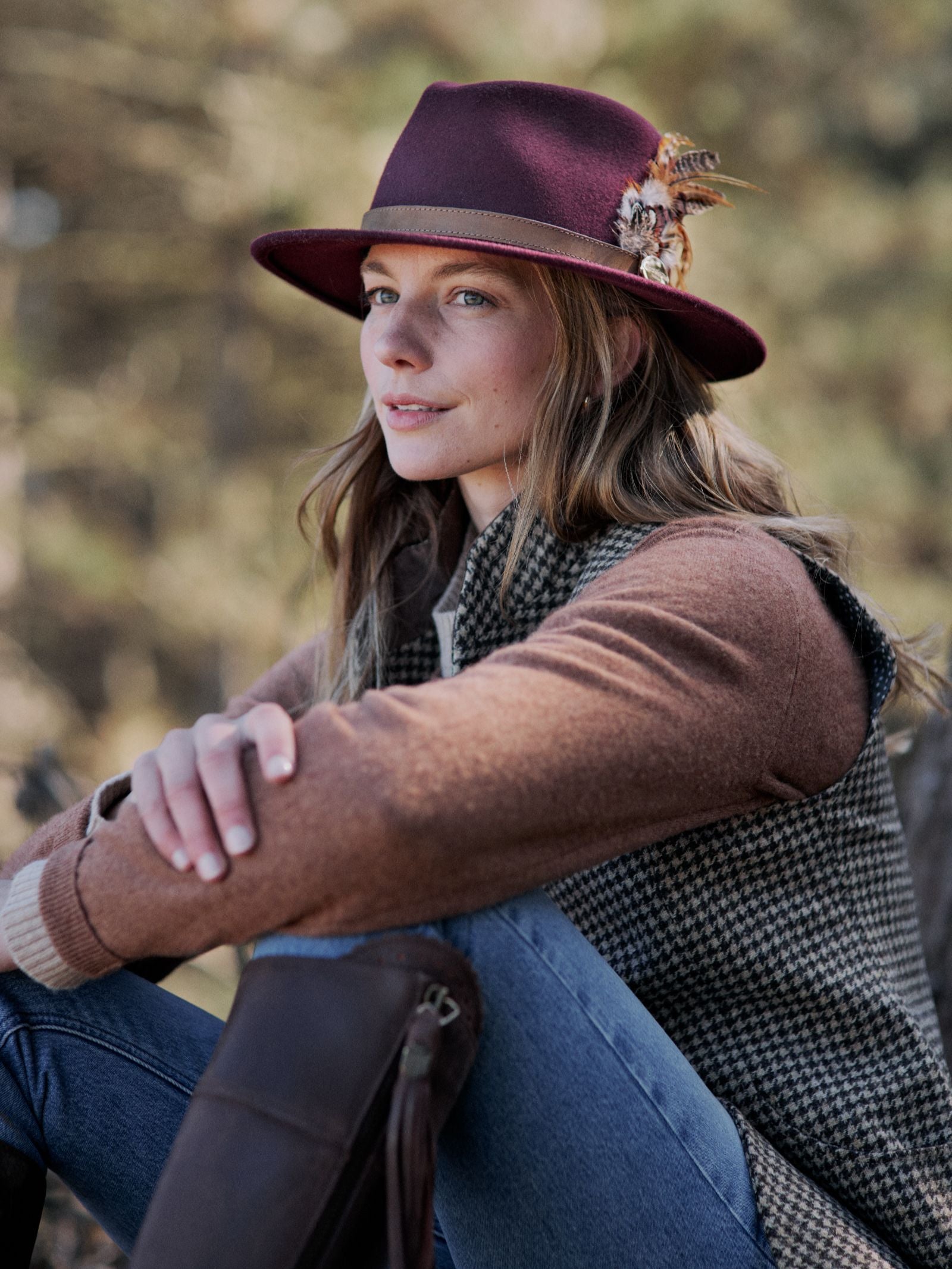 A woman in a Hicks & Brown Gamebird Fedora and a brown sweater with a houndstooth vest sits outdoors, one knee raised, with blurred trees in the background.