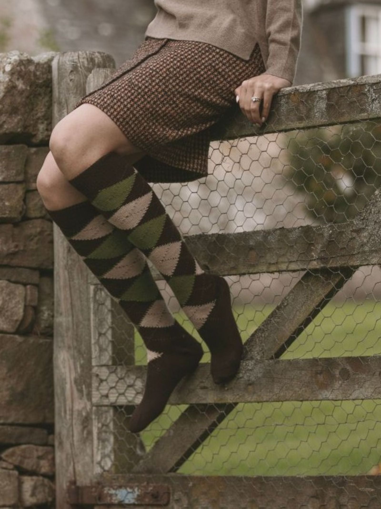 Wearing a brown plaid skirt, beige sweater, and Campbell’s of Beauly Argyle Sock handmade in Scotland, a person sits on a wooden gate with wire mesh in a rural outdoor setting. Their face is not visible.