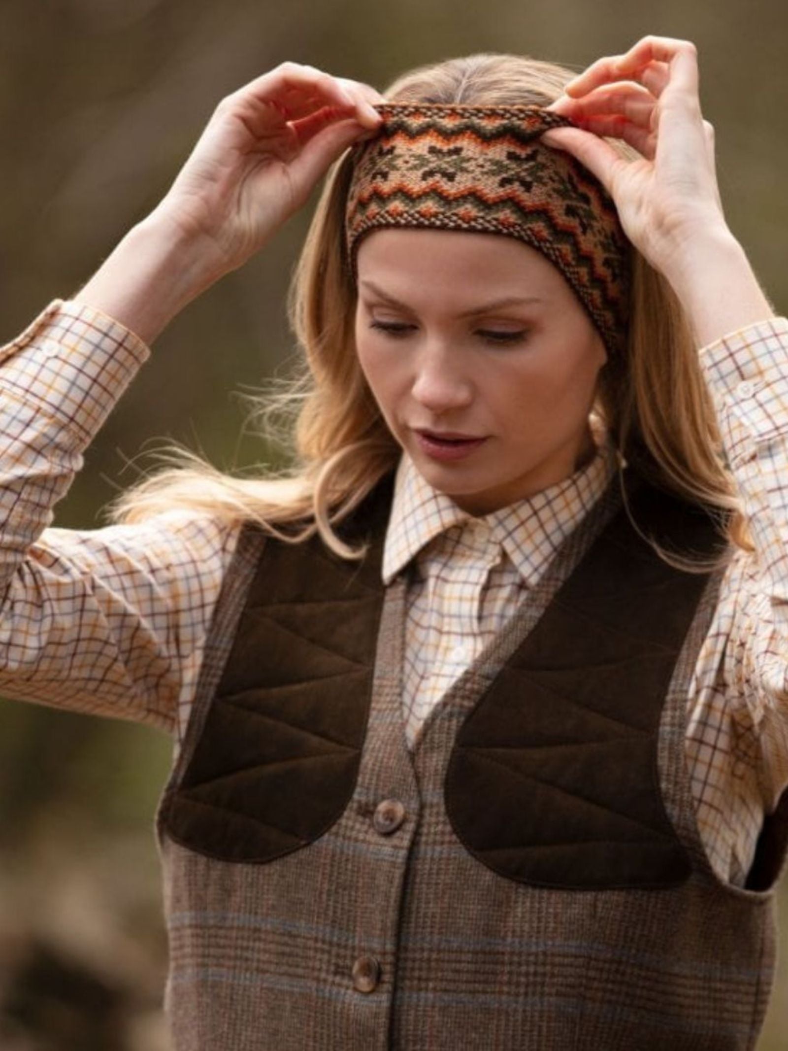 A woman in a brown plaid shirt and vest adjusts the Campbell's of Beauly Fairisle Headband, made from 100% wool in the Scottish Borders, while standing outdoors against a softly blurred background.
