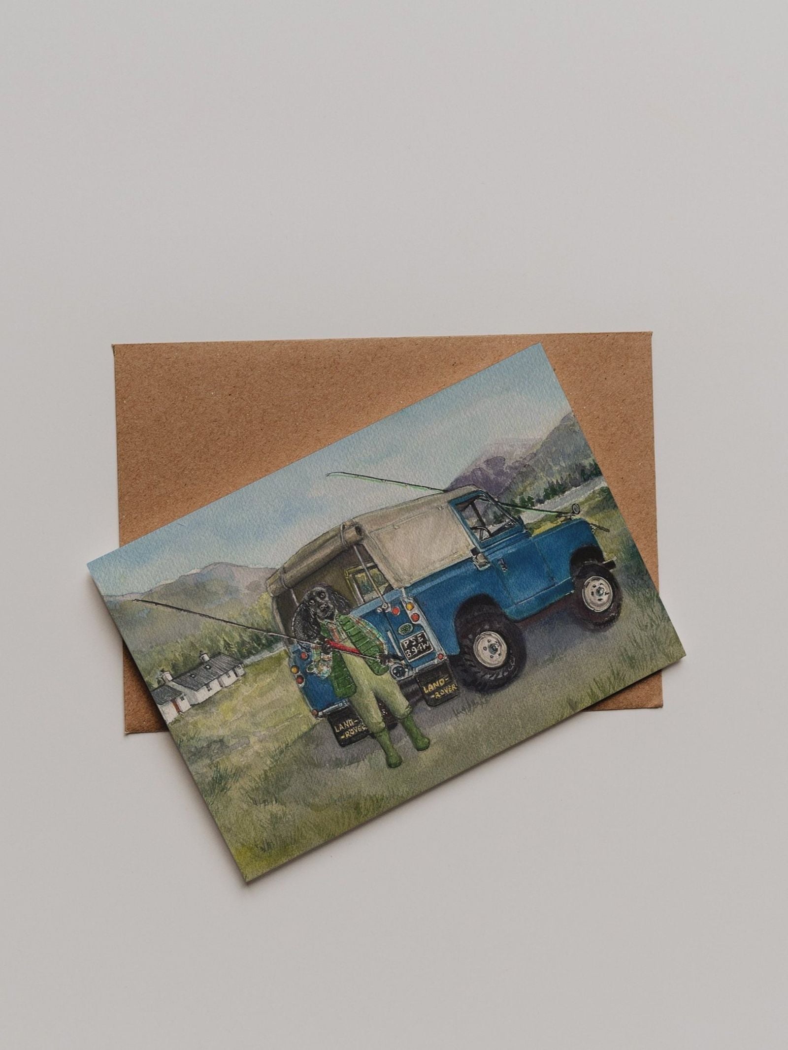 The Land Rover Cocker Greeting Card by Campbell’s of Beauly showcases an illustration of a blue off-road vehicle with a person in green standing beside it. Designed by Sophie Murray, the card is pictured resting on a brown envelope against a plain background.