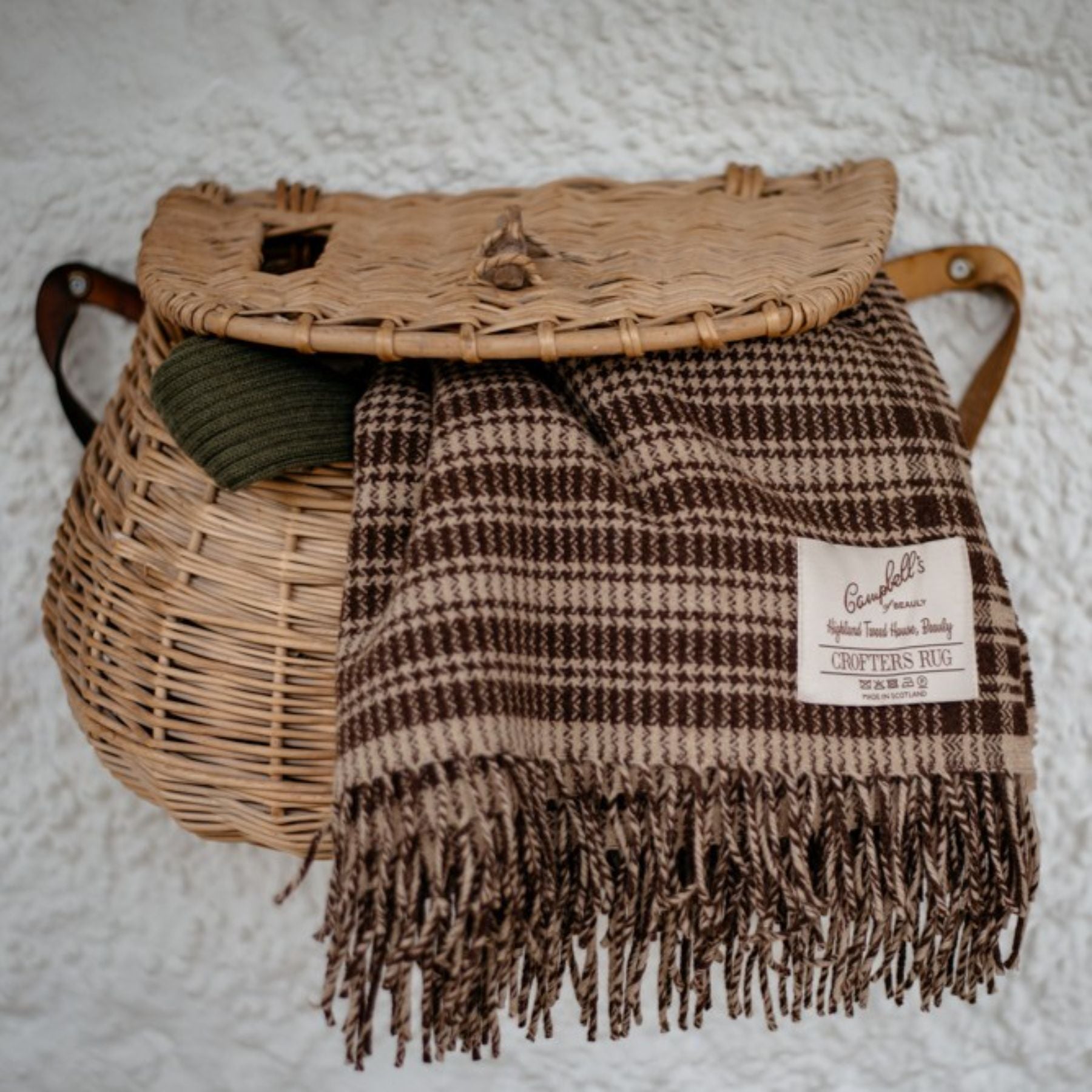 A woven basket with a lid hangs on a wall, with a brown checked wool blanket labeled Crofters Rug and a green knit item partially inside. The background is a textured white surface.