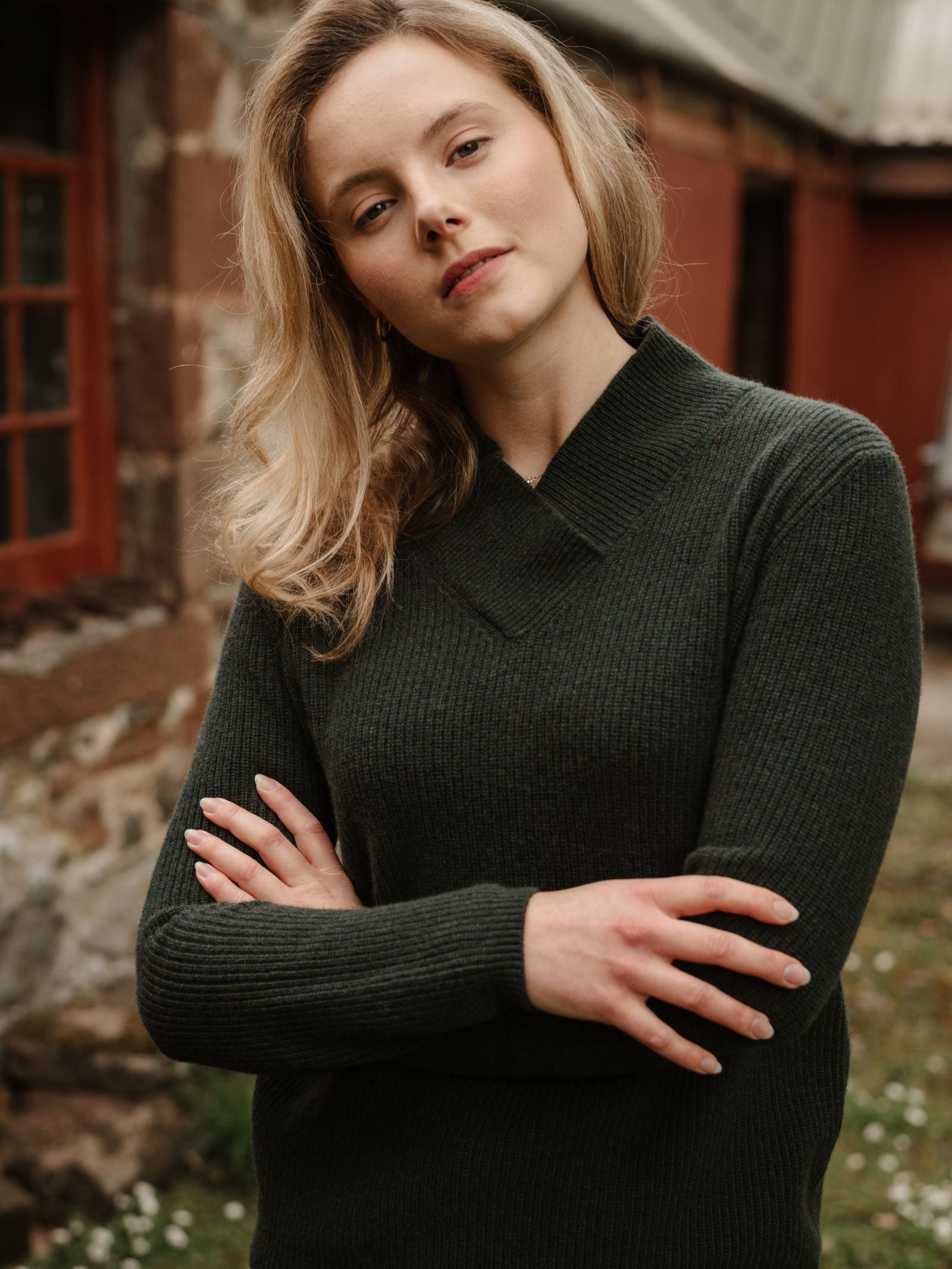 A woman with blonde hair stands outside with her arms crossed, confidently looking at the camera. She is wearing the Rib V Neck Jumper by Campbell’s of Beauly. In the background, there is a stone wall and a red building.