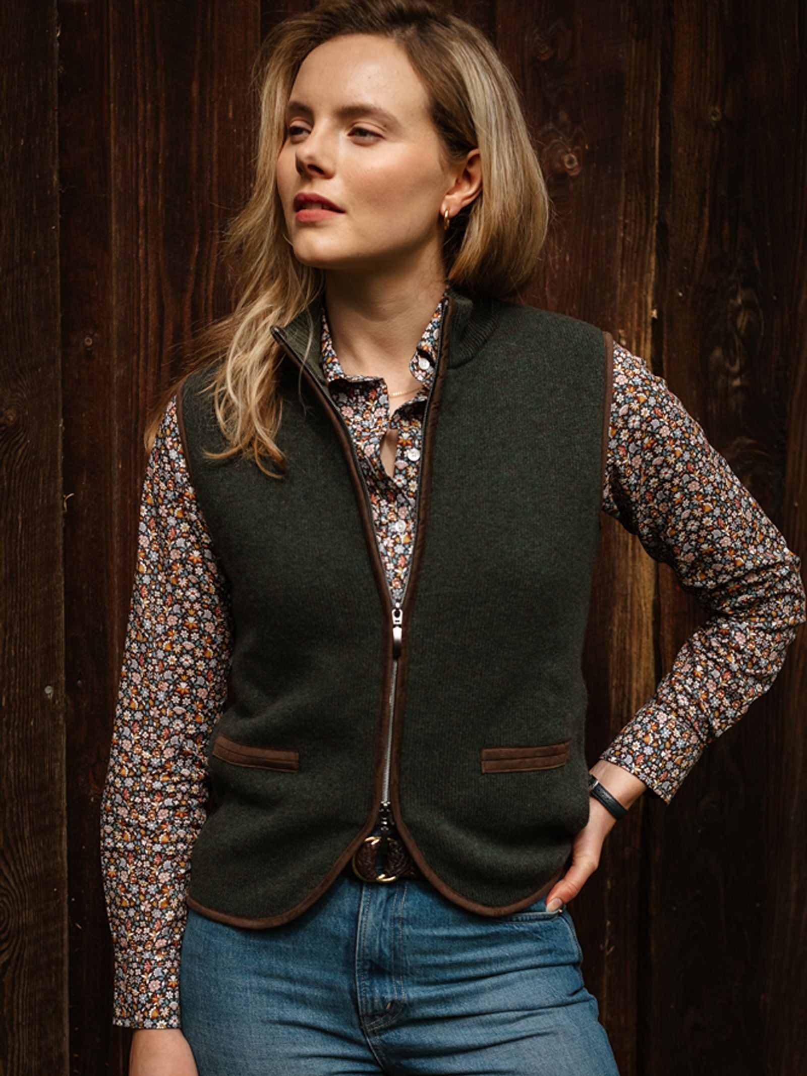 A woman stands in front of a wooden wall, looking to the side with one hand on her hip, effortlessly capturing timeless countryside style in a Campbell’s of Beauly Merino Cashmere Gilet layered over a floral shirt and blue jeans.