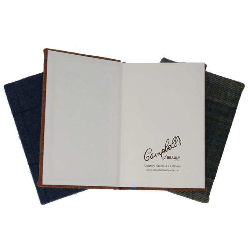 An open A5 Campbells of Beauly Tweed Notebook with a blank right page and branding on the left rests atop two closed notebooks featuring sustainable blue and green fabric covers.