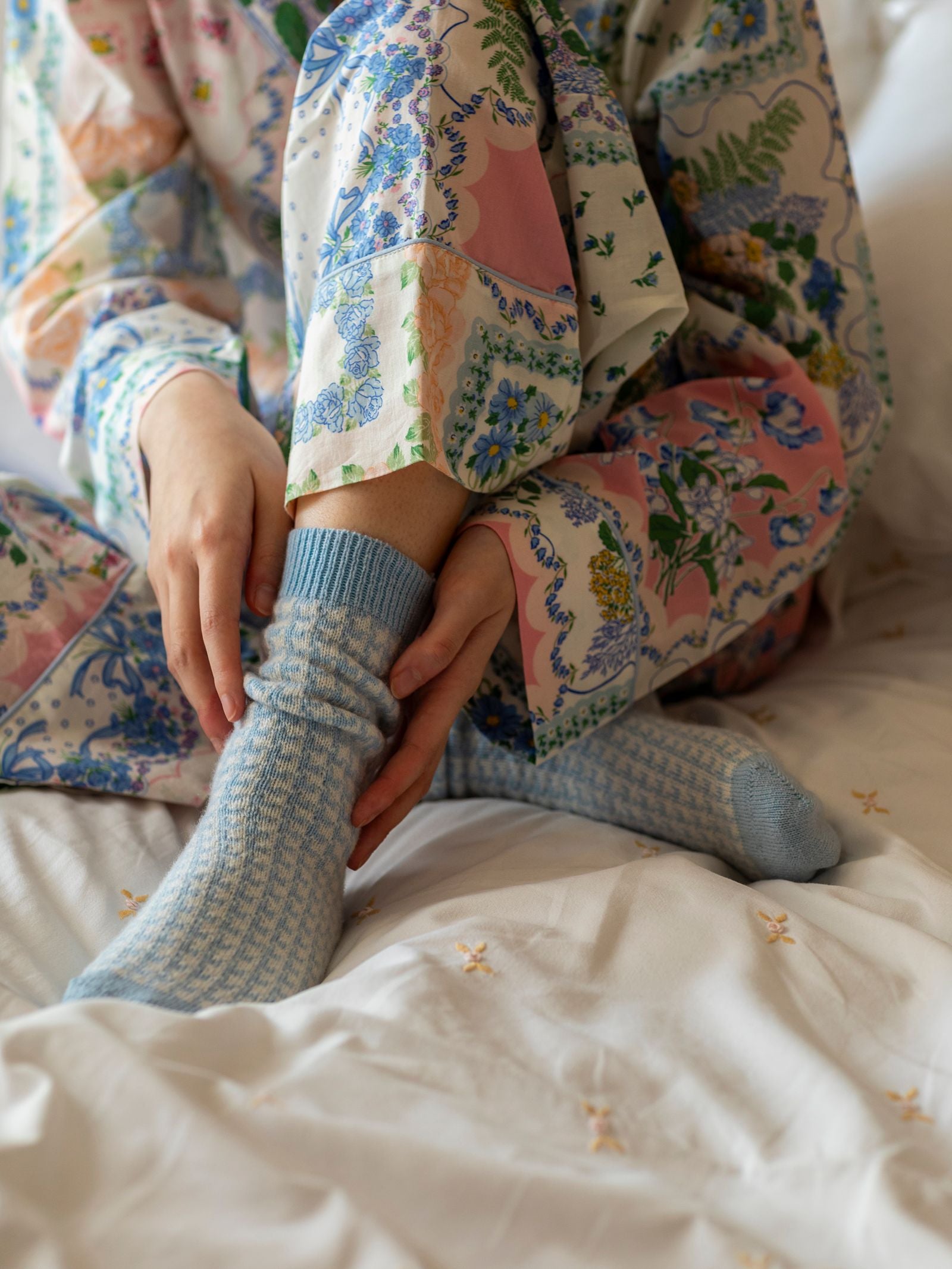 A person in colorful floral pajamas sits on a bed, adjusting their Campbell’s of Beauly Cashmere Houndstooth Socks. The bedding is white with small embroidered designs.
