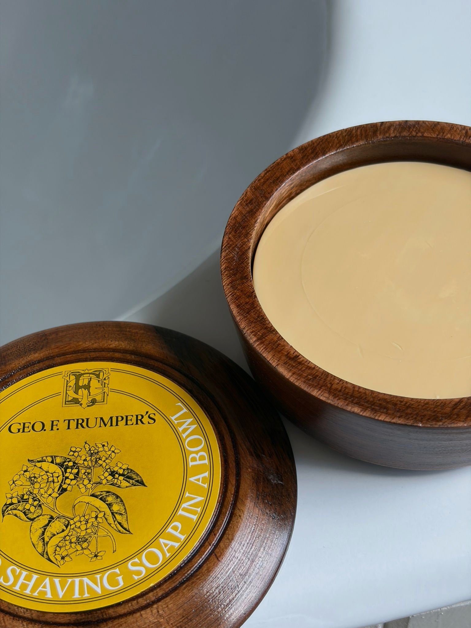 Geo. F. Trumper’s Sandalwood Shaving Soap & Bowl sits beside its matching wooden lid, featuring a yellow label and floral design—an elegant choice for a refined wet shaving experience.