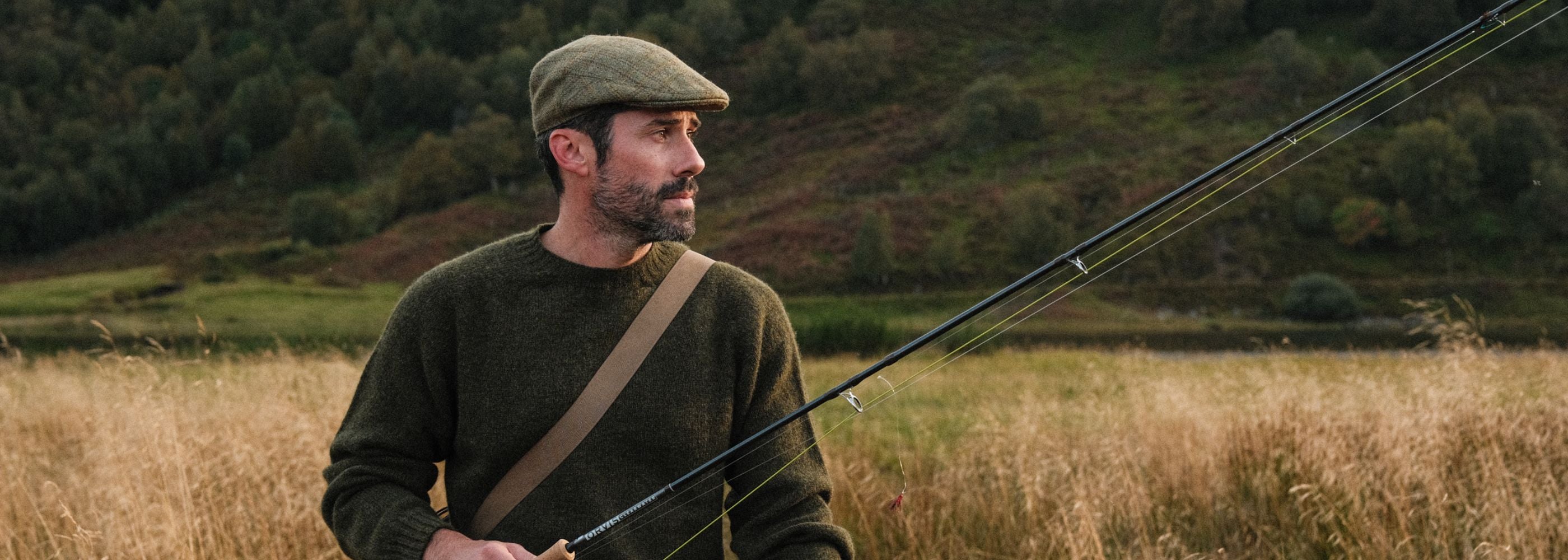 A man wearing a flat cap, green sweater, and tan shoulder strap holds a fishing rod while standing in a grassy field with a forested hillside in the background.