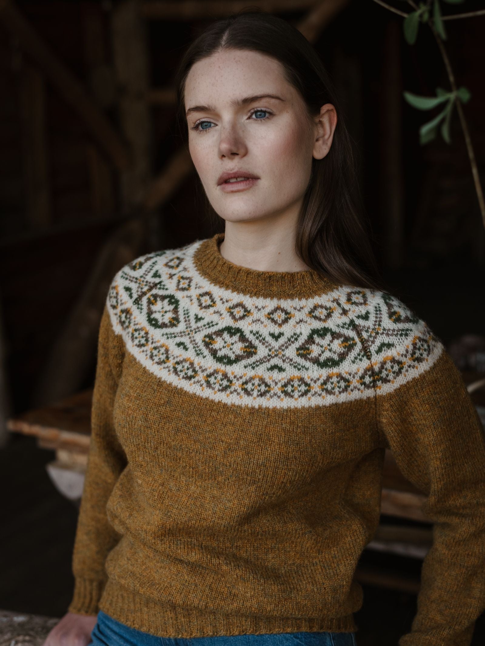 A woman with long brown hair wears the Campbell's of Beauly Shetland Fairisle Crew, a mustard yellow jumper with a white, green, and brown geometric yoke. She stands indoors, looking thoughtful.