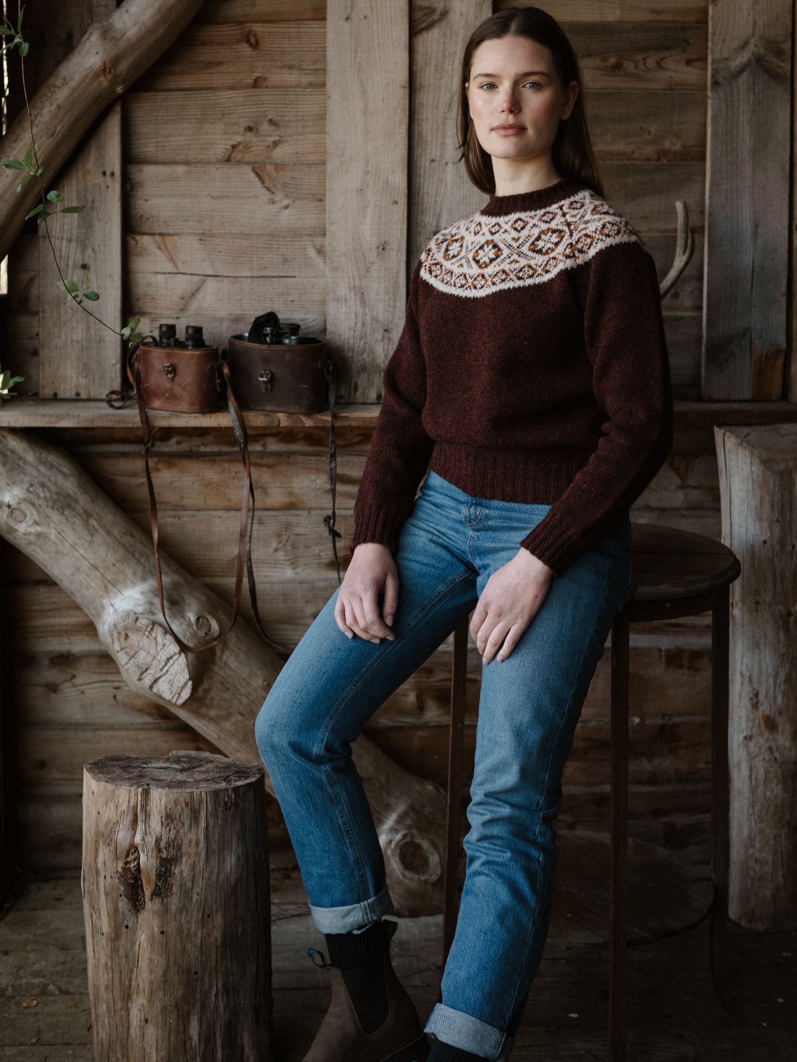 A woman with straight brown hair sits on a stool in a rustic cabin, wearing Campbell’s of Beauly’s Shetland Fairisle Crew in maroon, blue jeans, and brown boots. Vintage cameras hang on the wooden wall behind her.