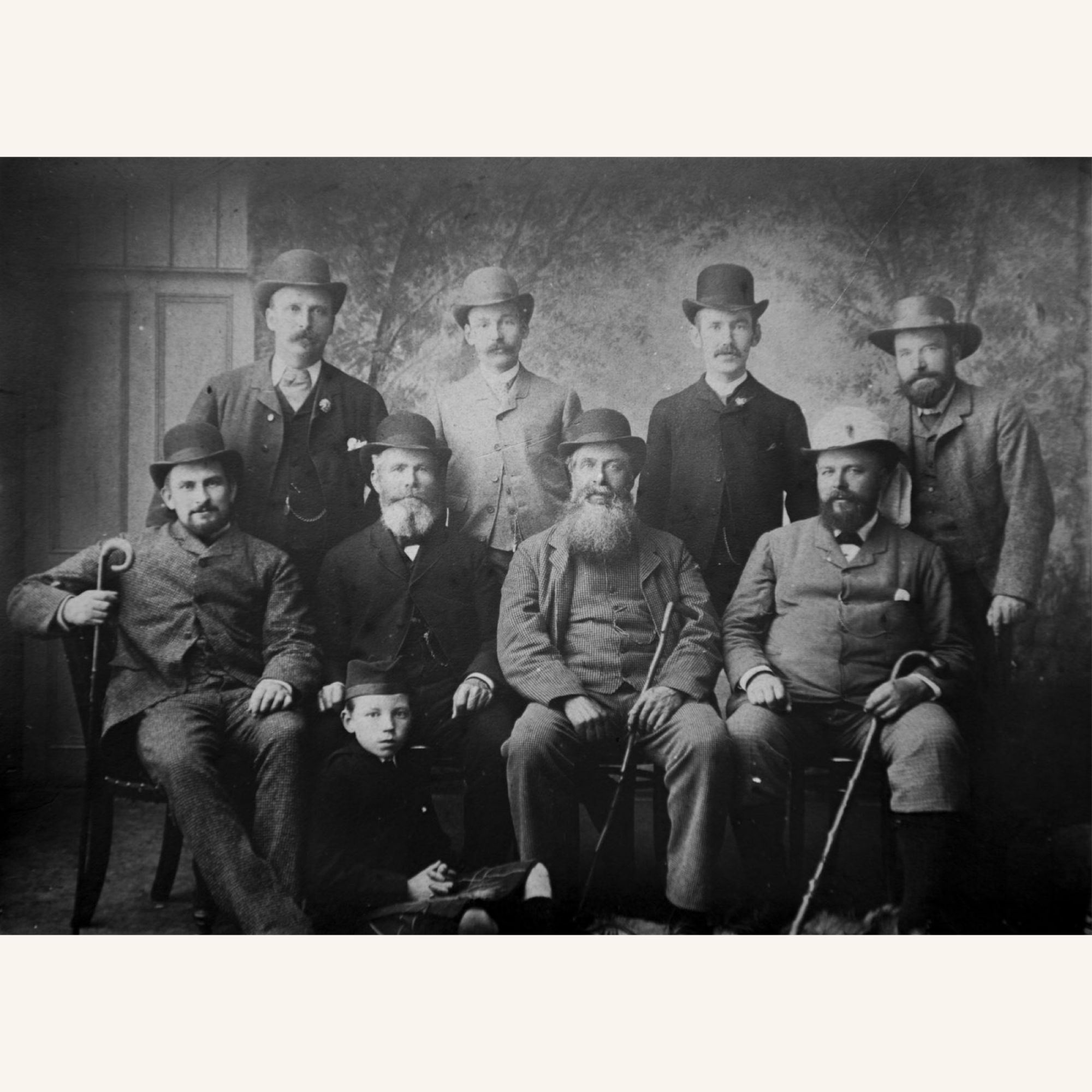A black and white vintage photo of nine men and one boy, all wearing hats and suits. The men sit and stand in two rows, some holding canes, in front of a painted backdrop. The boy sits on the floor in front.