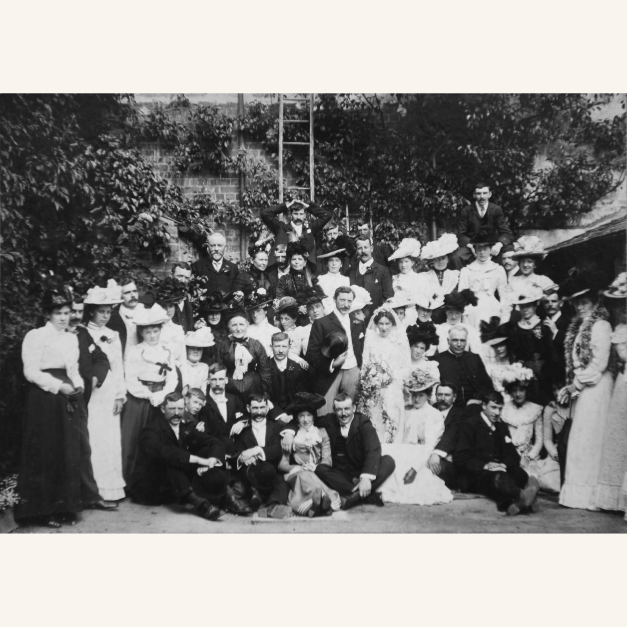 A large group of men, women, and children in formal Victorian-era clothing from Campbells of Beauly pose for a black-and-white outdoor group portrait, likely at a wedding, with greenery and a brick wall in the background.