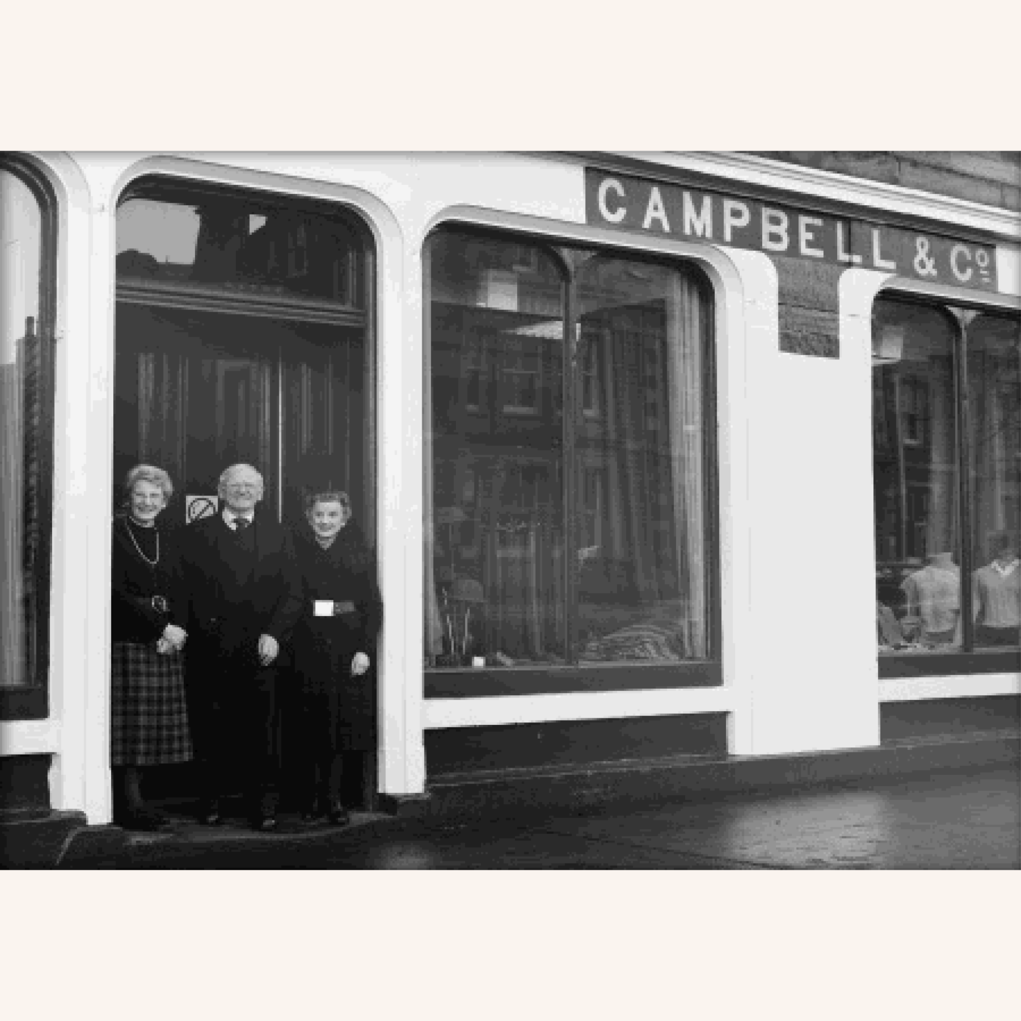 Three people stand in front of a storefront with large windows and the sign CAMPBELL & CO above the entrance. The shop windows display clothing inside. The image is in black and white.