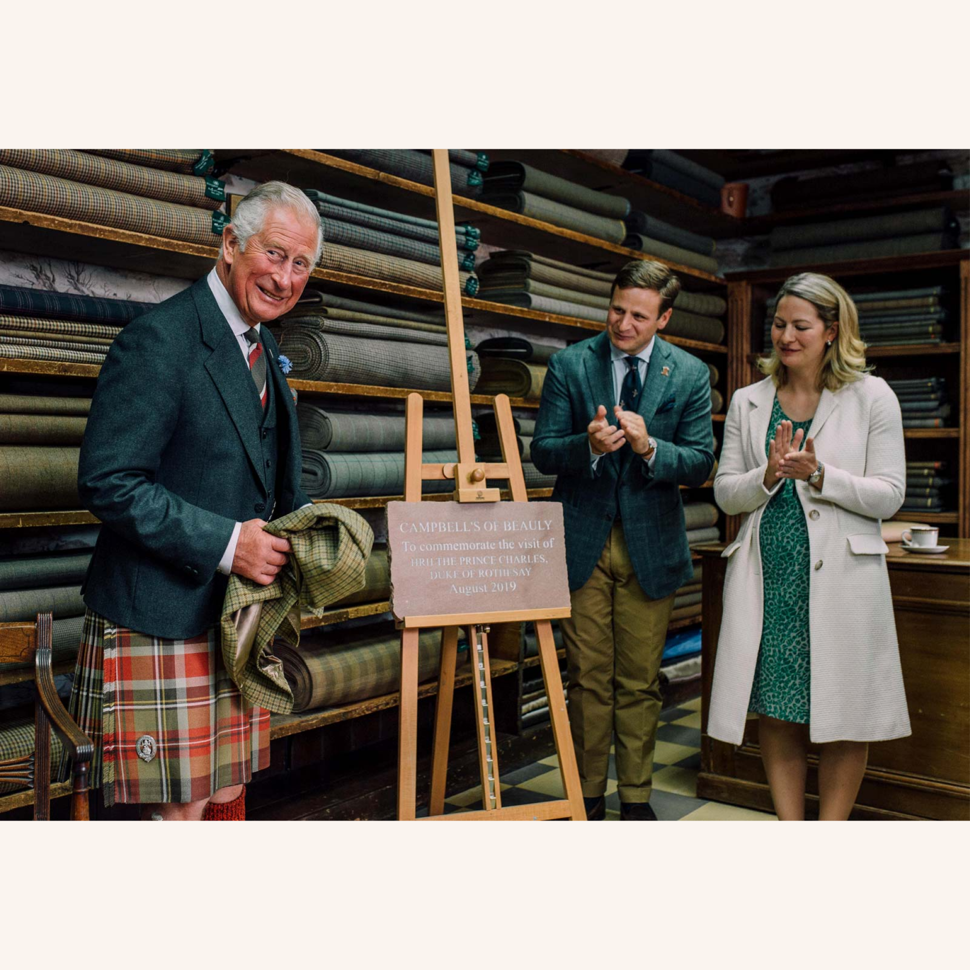 A smiling man in a plaid kilt unveils a plaque in a fabric shop, while a man and woman beside him clap and smile. Rolls of patterned fabric are stacked on shelves in the background.