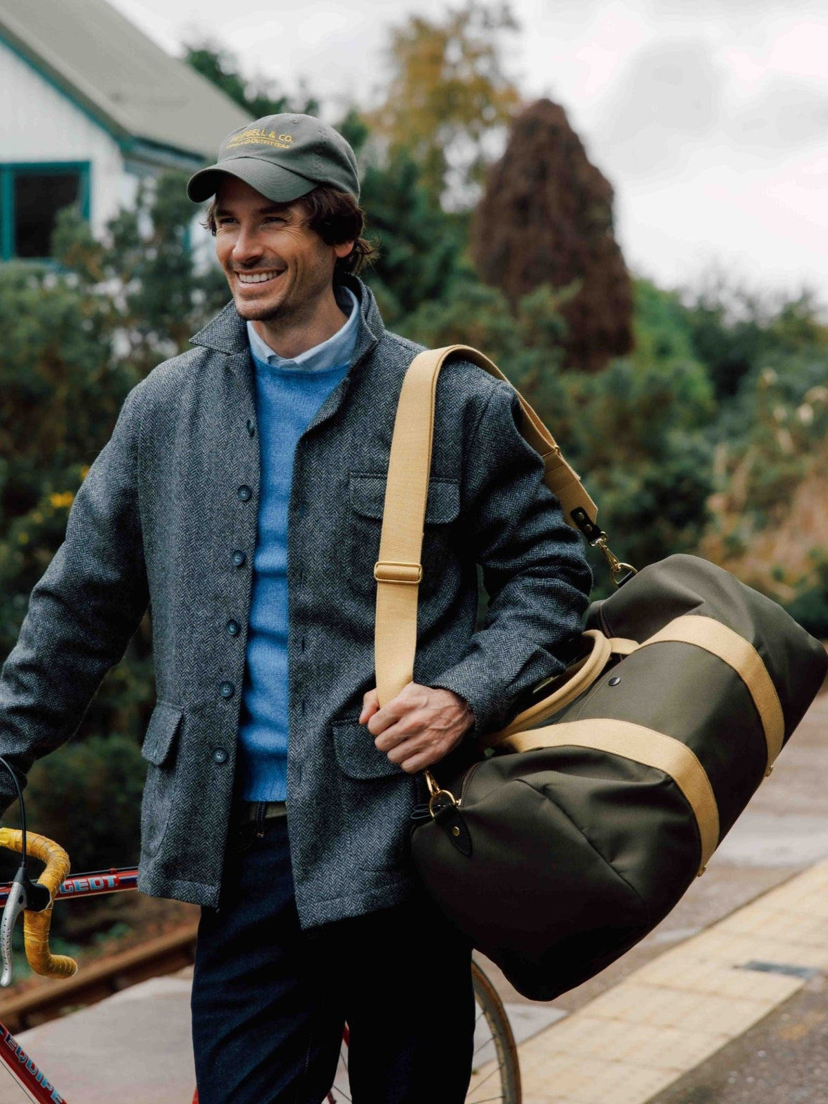 A smiling man in a gray jacket and cap carries the Campbell’s of Beauly Utility Medium Duffle—a large green bag with tan straps—on his shoulder and holds a bicycle while outdoors near a house and greenery.