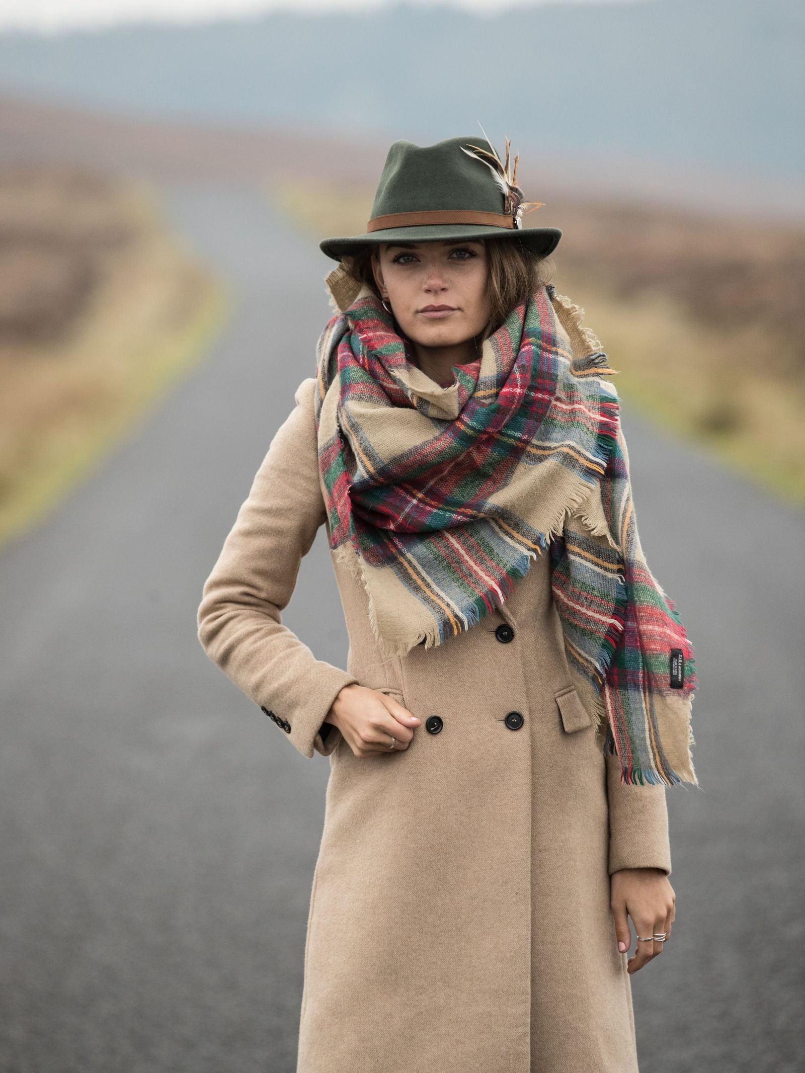 A woman in a tan coat, plaid scarf, and a green Hicks & Brown Gamebird Fedora stands on an empty rural road with soft, blurred hills in the background.