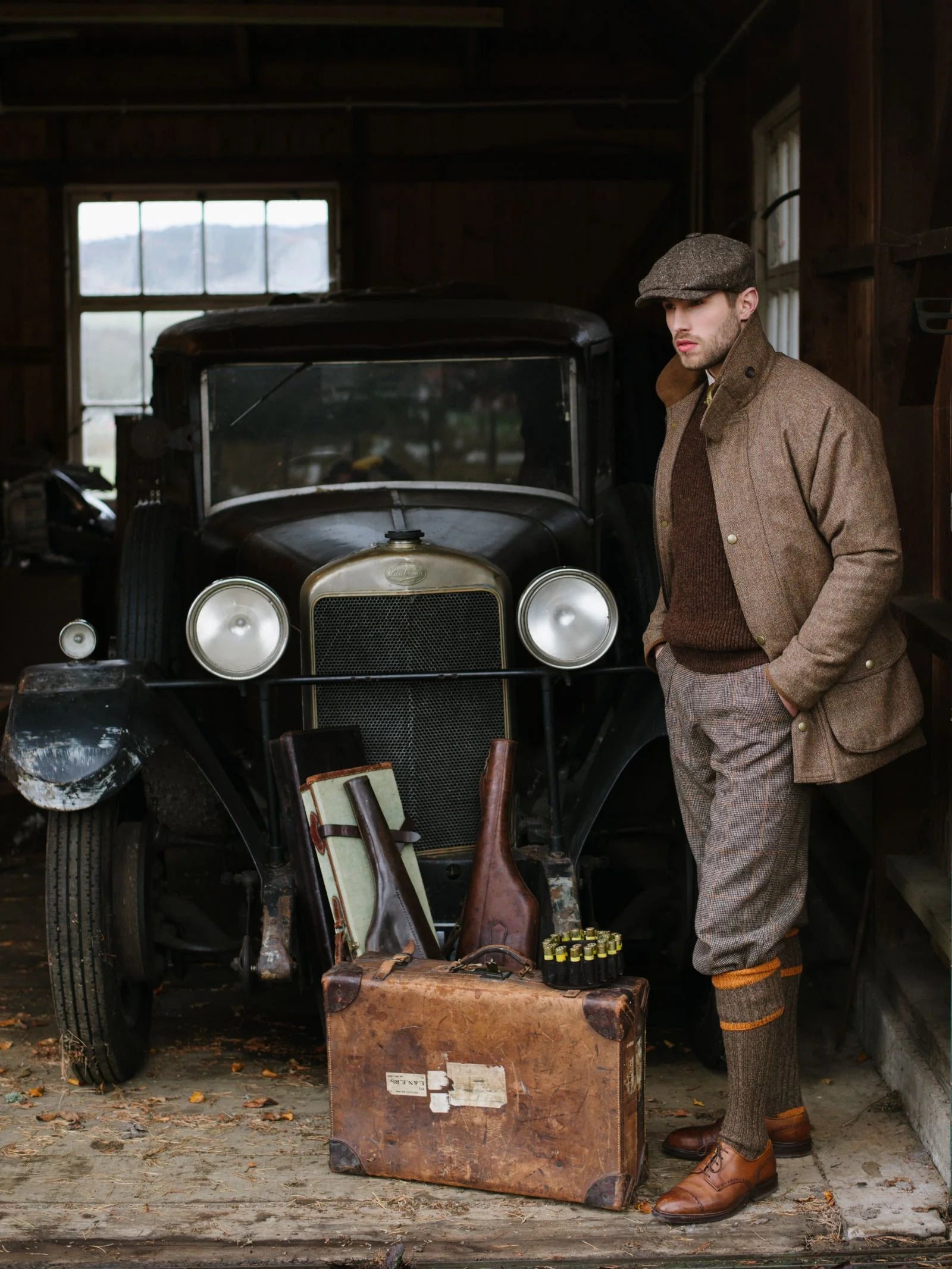 A man wearing Campbells of Beauly Plus Fours stands beside a vintage car in a dim garage, with a suitcase, two rifles, and shotgun shells laid out in front of the vehicle.