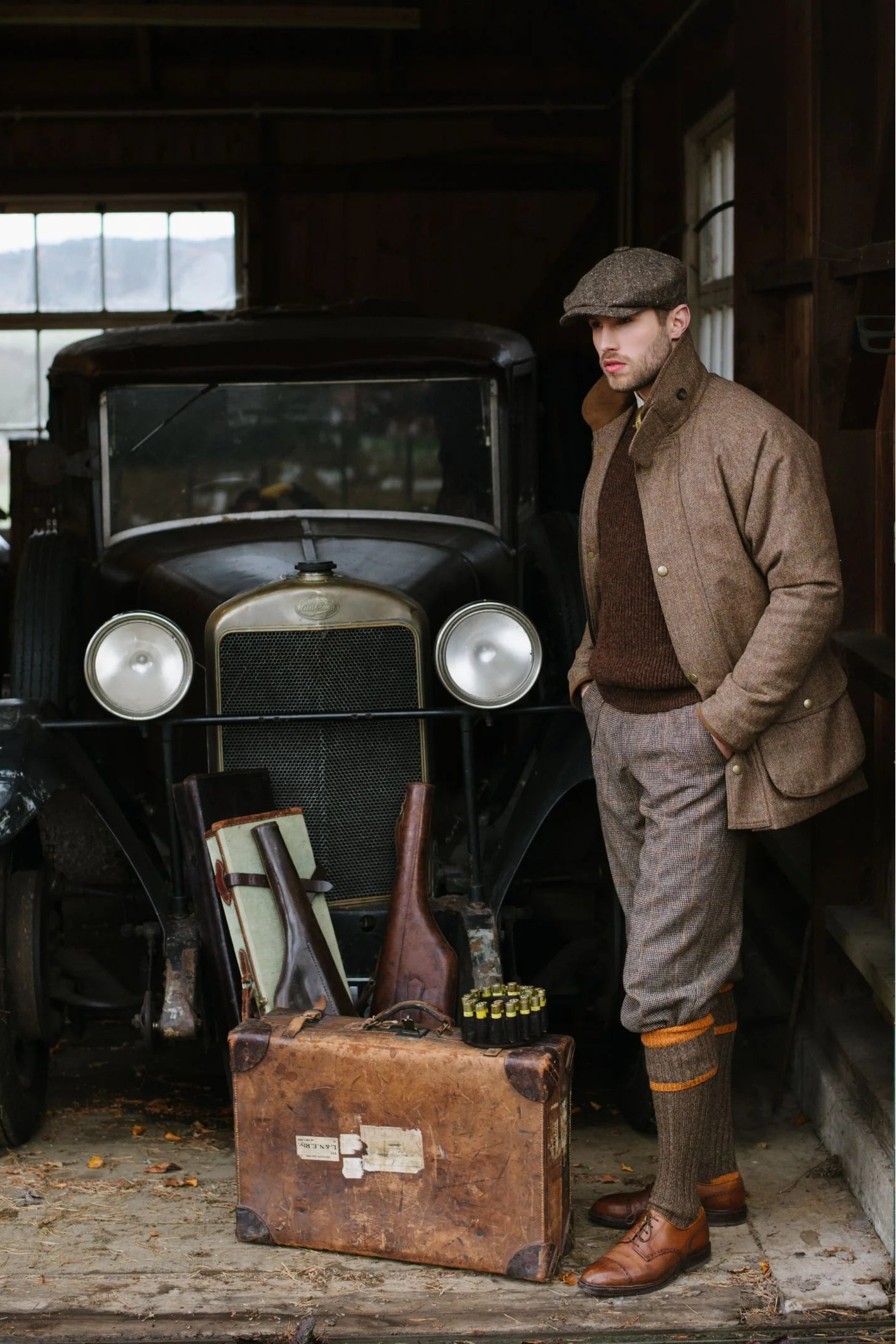 A man wearing Campbells of Beauly Plus Fours stands beside a vintage car in a dim garage, with a suitcase, two rifles, and shotgun shells laid out in front of the vehicle.