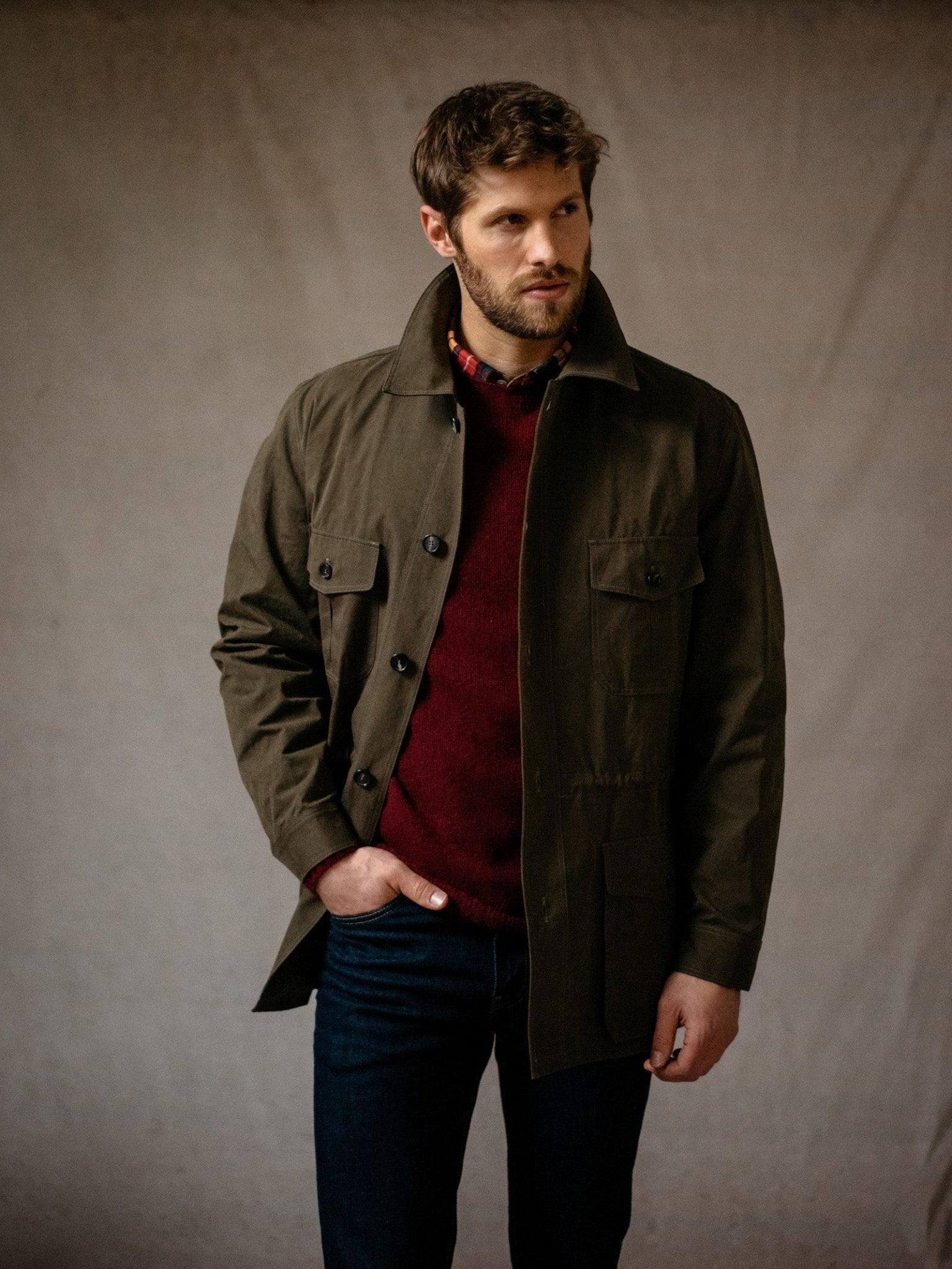 A bearded man stands with one hand in his pocket against a neutral backdrop, wearing the Campbells of Beauly Aigas Field Jacket in dark green with a water-repellent finish over a burgundy sweater and blue jeans.