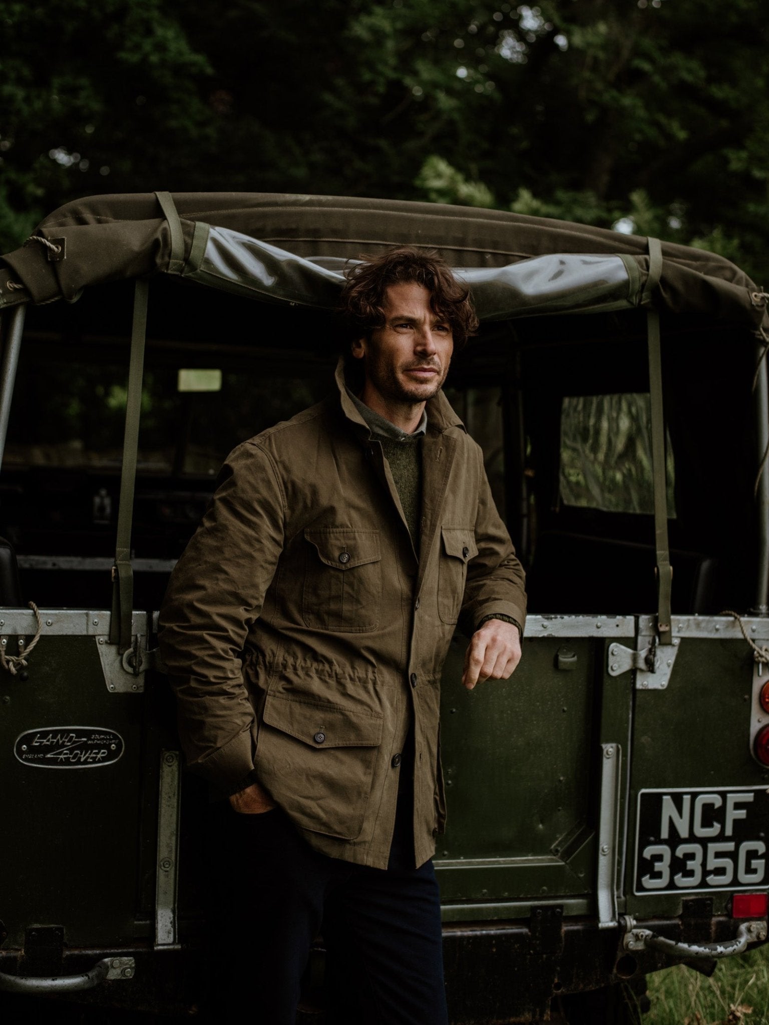 A man with wavy hair, dressed in Campbells of Beauly Aigas Field Jacket in brown and dark pants, leans casually against a green vintage Land Rover amid a wooded outdoor scene.
