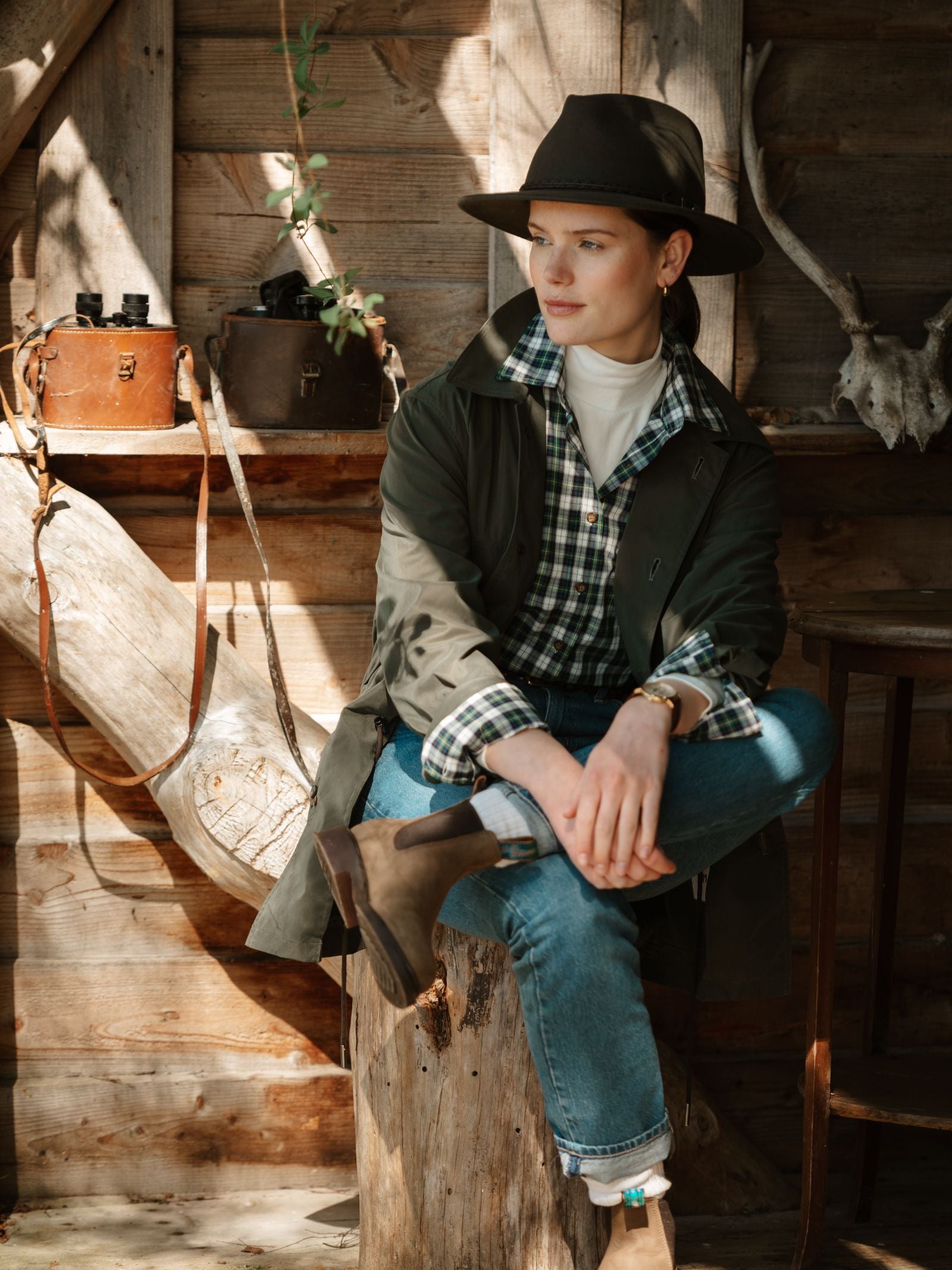 A person in an Akubra Unisex Traveller Hat sits indoors on a tree stump, surrounded by rustic wooden walls, vintage cameras, and antlers as sunlight streams in, creating a warm countryside vibe.