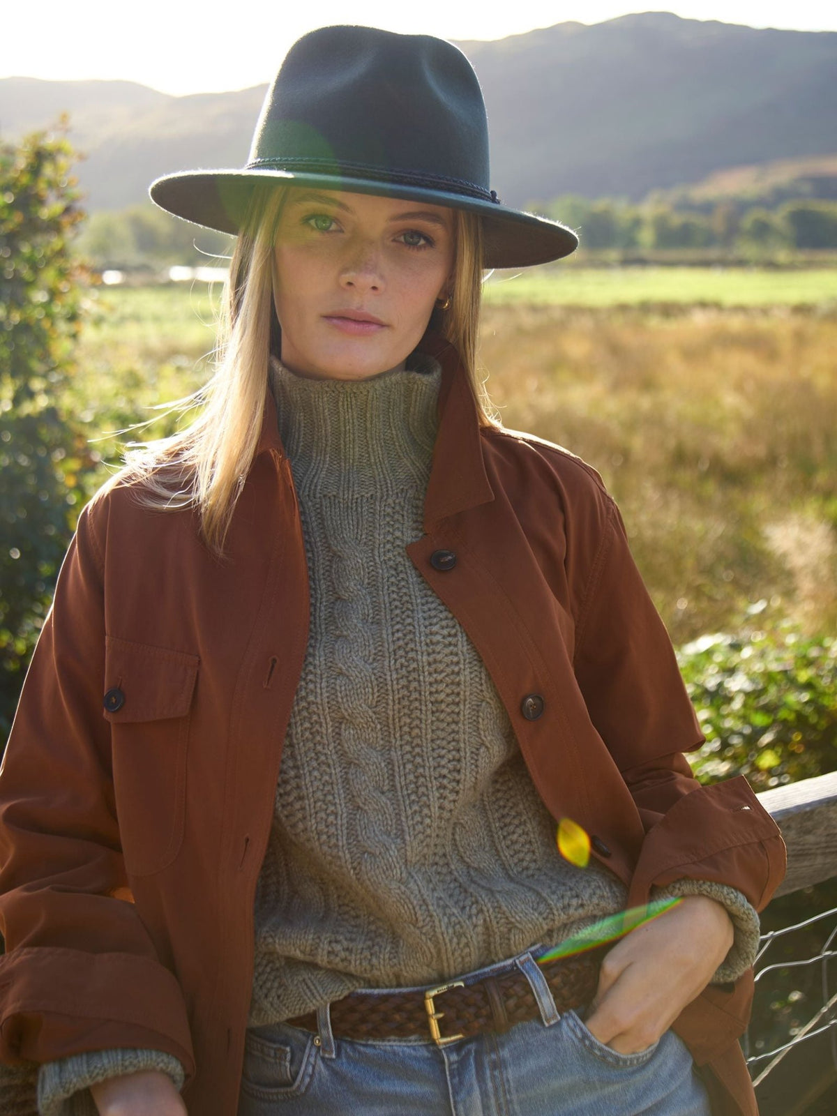 A woman wearing a supersoft Campbell’s of Beauly Cable Roll Neck Jumper, a rust-colored jacket, and a wide-brimmed hat standing outdoors.