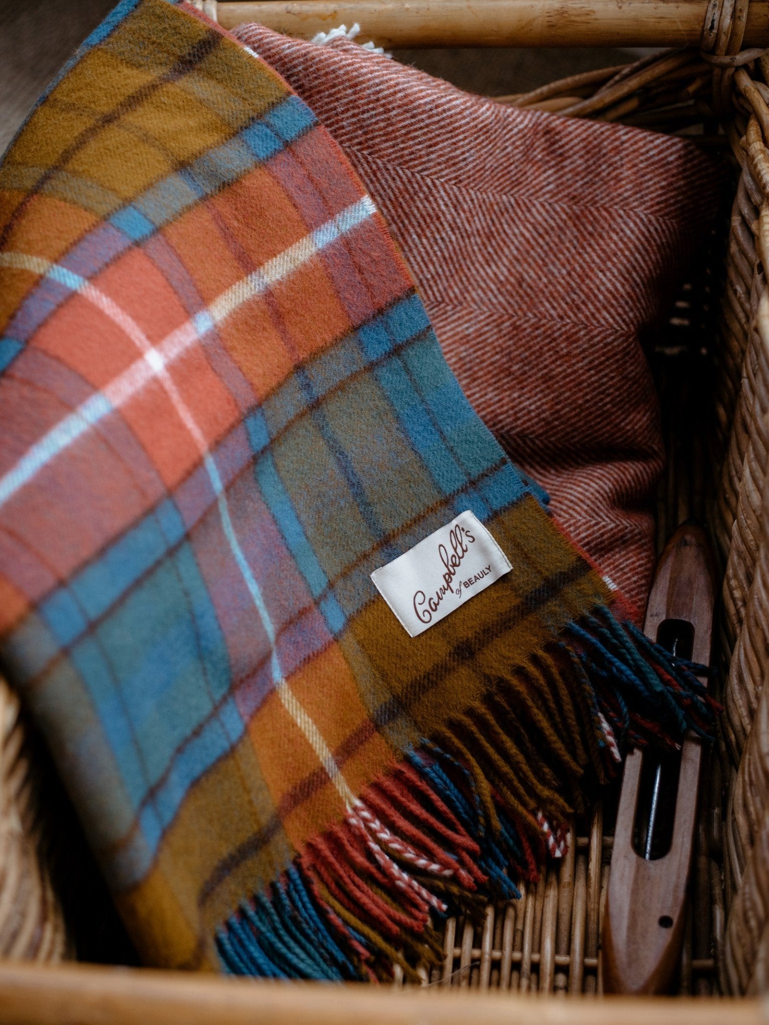 A woven basket contains a colorful plaid wool scarf labeled Campbells of Beauly, a red herringbone textile, an Antique Buchanan Tartan Lambswool Blanket by Campbells of Beauly, and a wooden weaving shuttle.