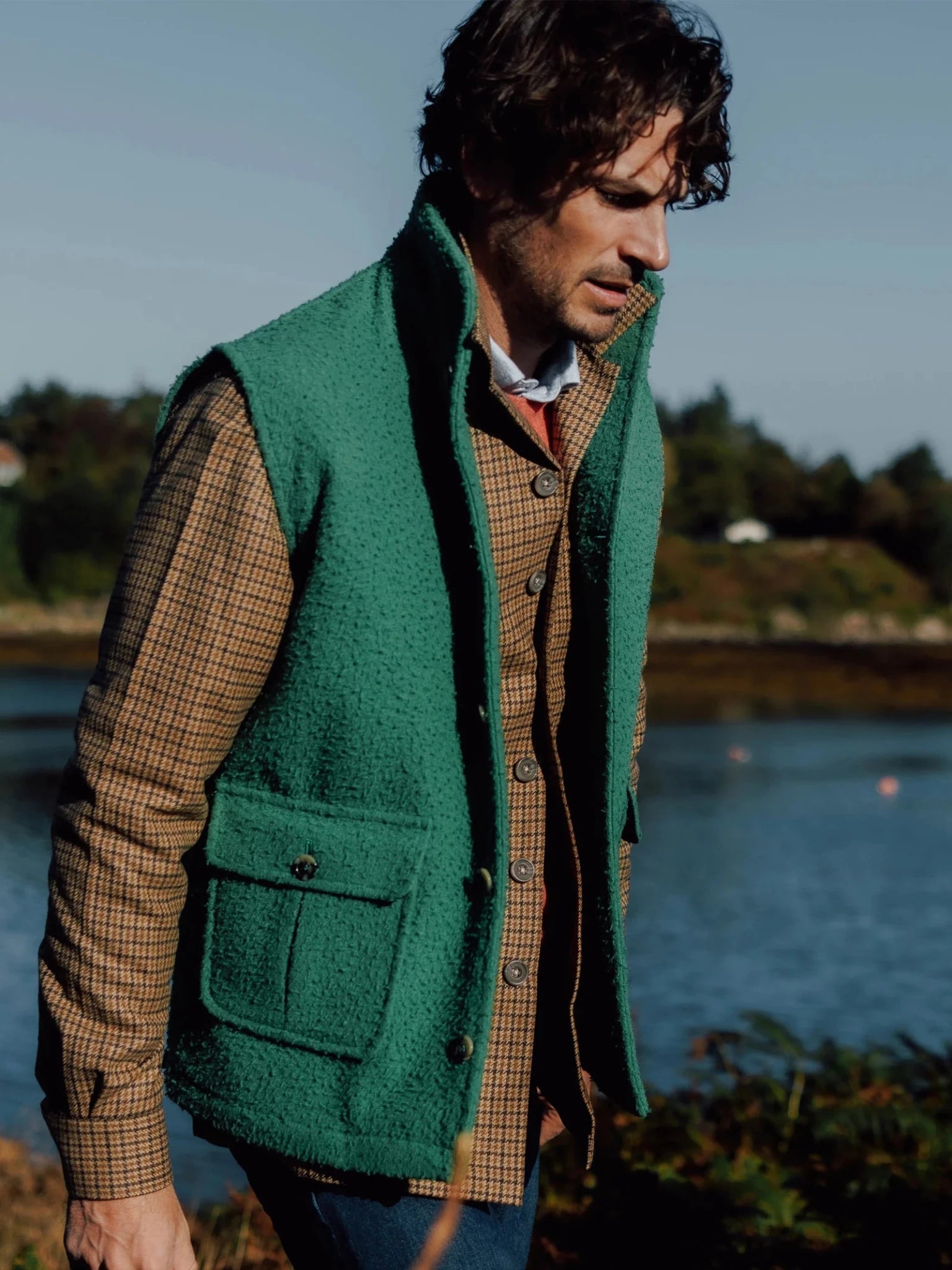 A man with wavy dark hair wears the Auld Stock Casentino Fleece Gilet in Lichen from Campbell's of Beauly over a brown plaid shirt, standing outdoors by water with trees and a blue sky in the background.