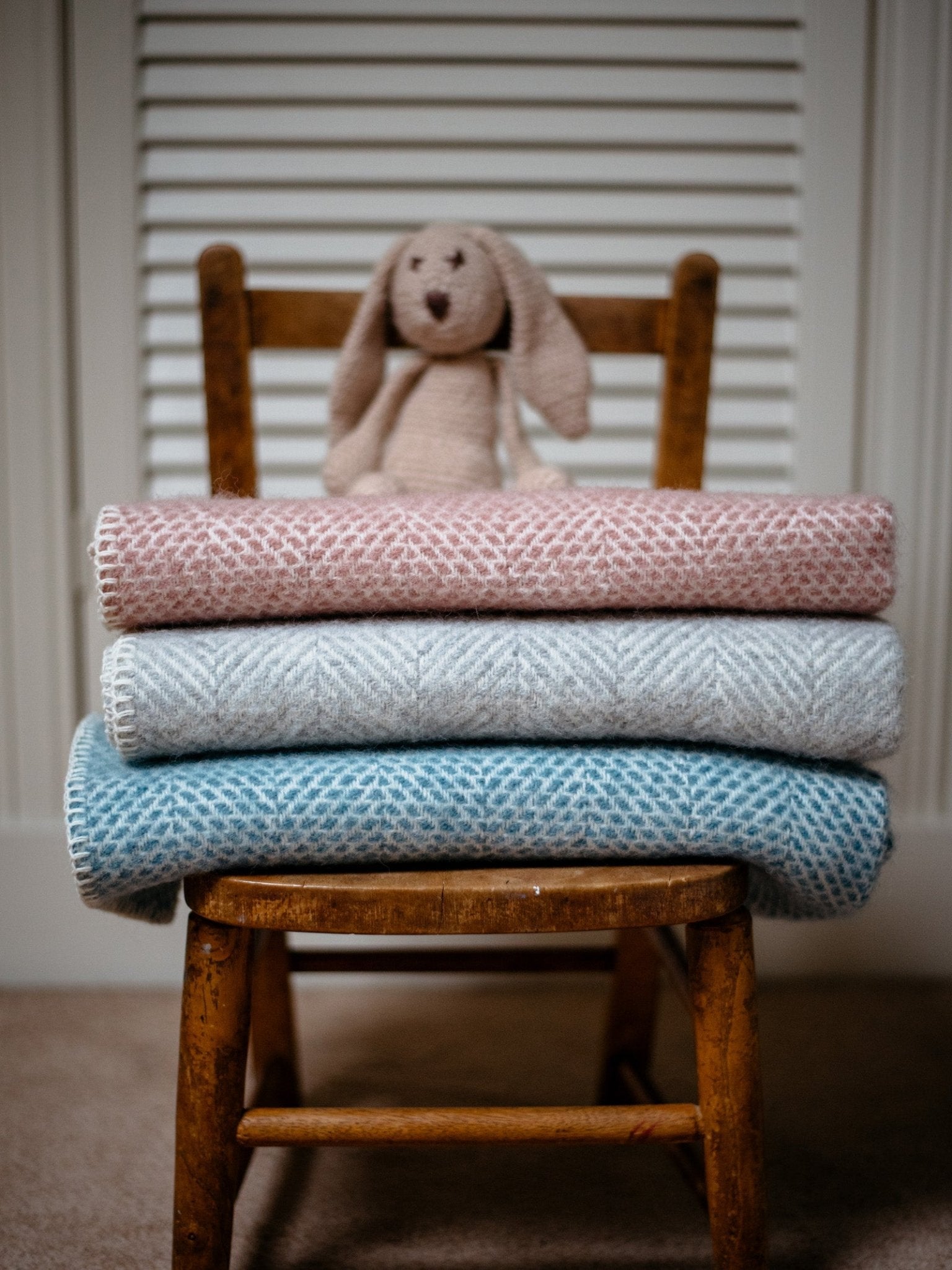 A wooden chair holds three Campbells of Beauly Baby Blankets—pink, gray, and blue—in a soft weave for year-round comfort. A plush bunny sits on top, set before white louvered doors.