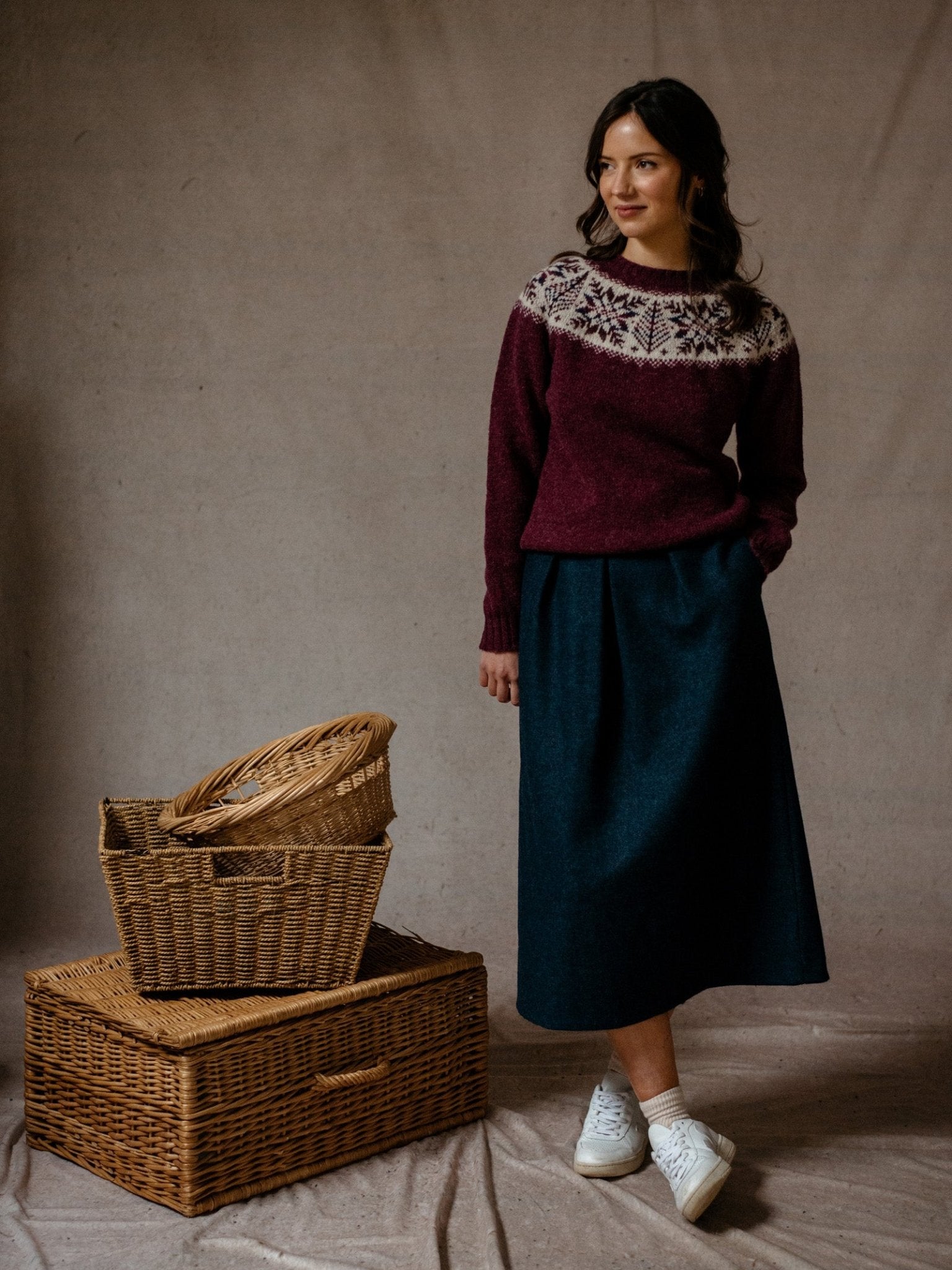 A woman stands indoors on a beige backdrop, wearing Campbells of Beauly Bell Skirt in Midnight Blue Tweed, a maroon sweater with a white and black Nordic pattern, and white sneakers. She poses with one hand in her pocket beside stacked wicker baskets.