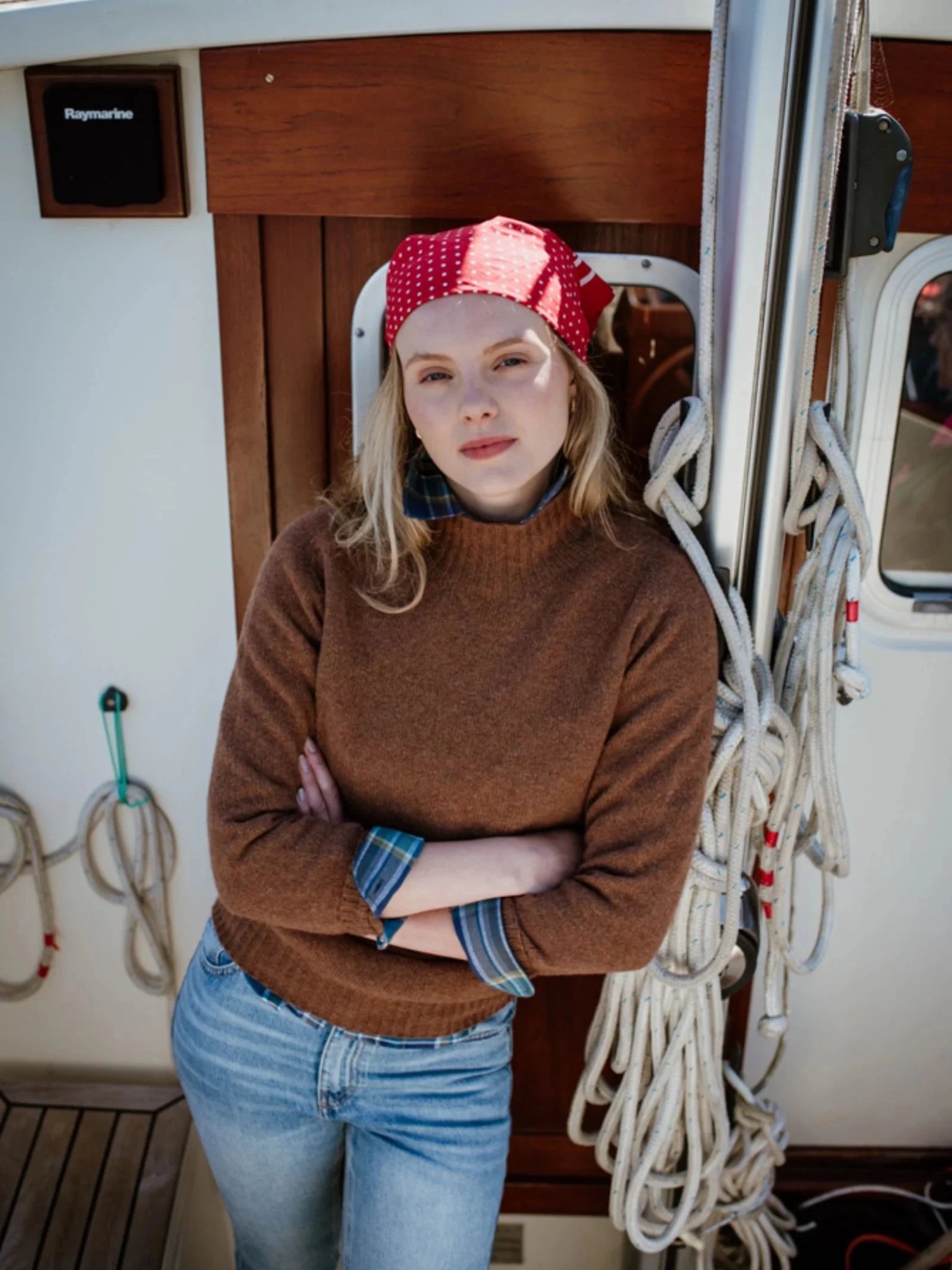 A woman in a red polka dot headscarf, Campbell's of Beauly Auld Stock Geelong Lambswool Funnel Neck Jumper, and jeans stands on a boat by coiled ropes, arms crossed and looking confidently at the camera.